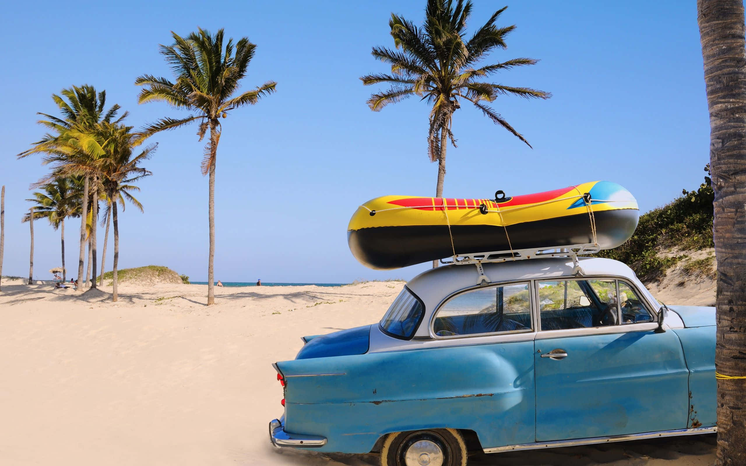 Beach Camping 桌布 壁紙 — 一輛復古藍色汽車，車頂綁著色彩繽紛的充氣筏，停泊在沙灘上，周圍是高高的棕櫚樹。