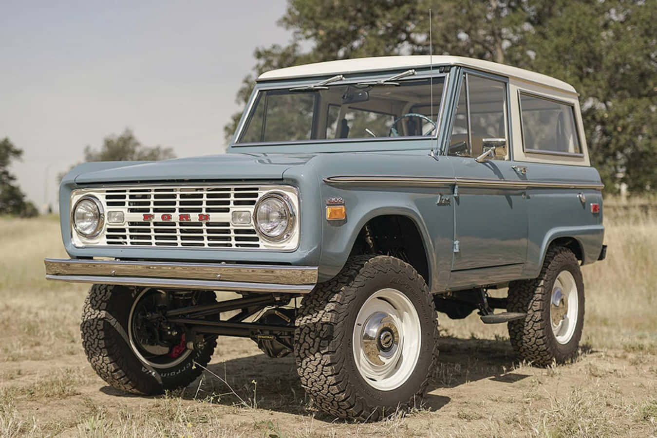 Ford Bronco 桌布 壁紙 — 一輛復古藍色福特Bronco，白色車頂和超大輪胎，在明亮的日光下自豪地矗立在乾燥的草地上。