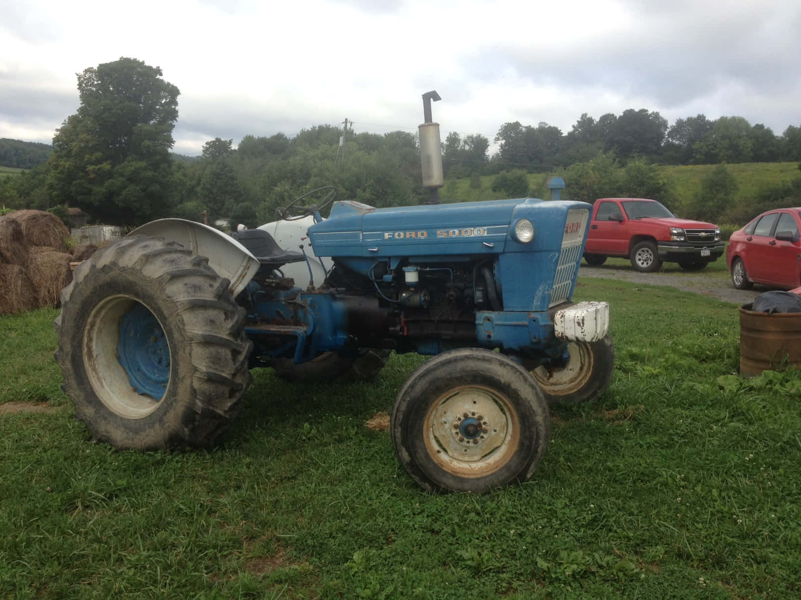 Tractor 桌布 壁紙 — 一輛復古藍色福特拖拉機停泊在草地上，旁邊有乾草捆，背景有紅色汽車和樹木，天空多雲。