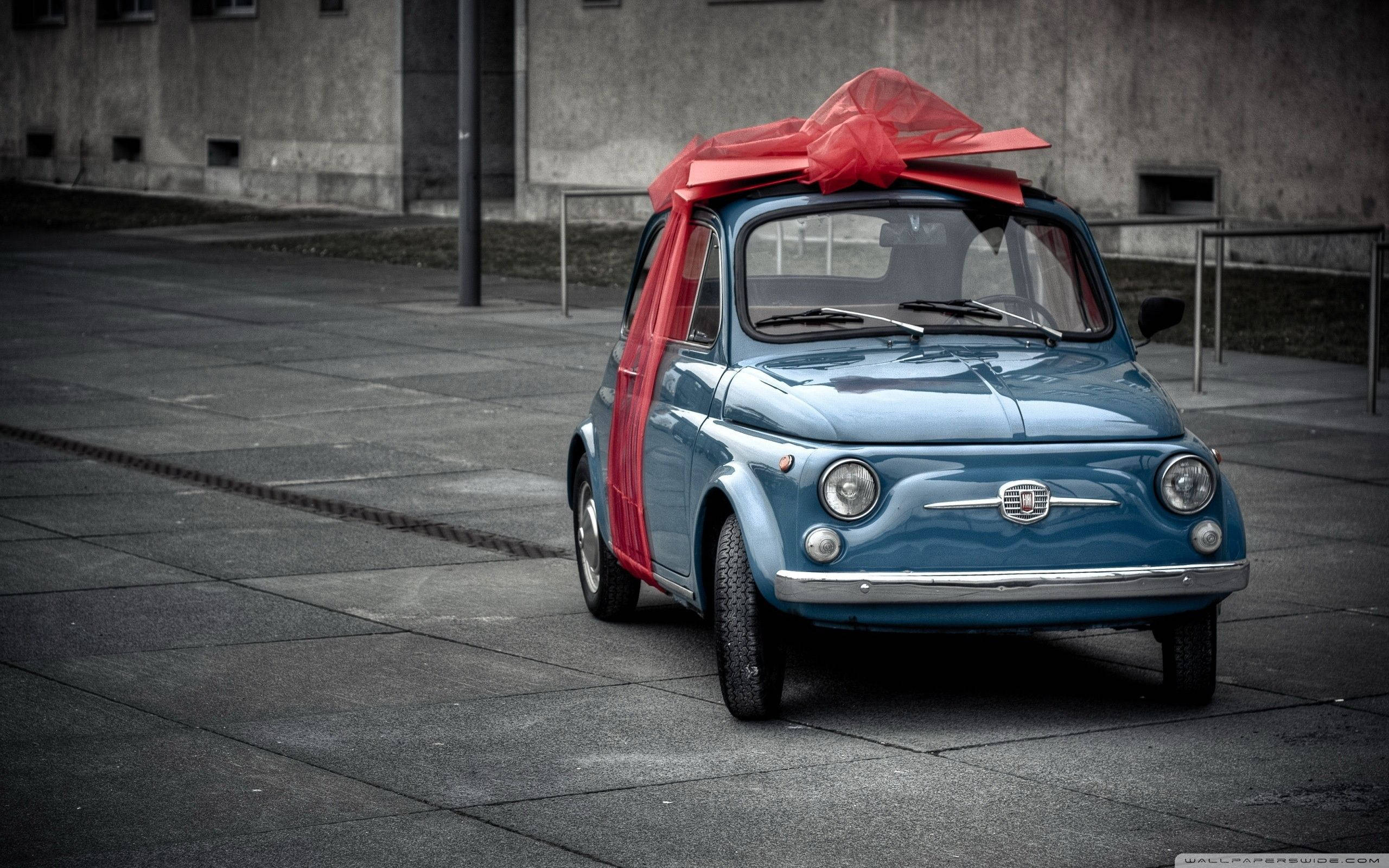 Fiat 桌布 壁紙 — 一輛復古藍色飛雅特500，車頂裝飾著一個巨大的紅色蝴蝶結，停在灰色混凝土廣場上，背景是風化的牆壁。