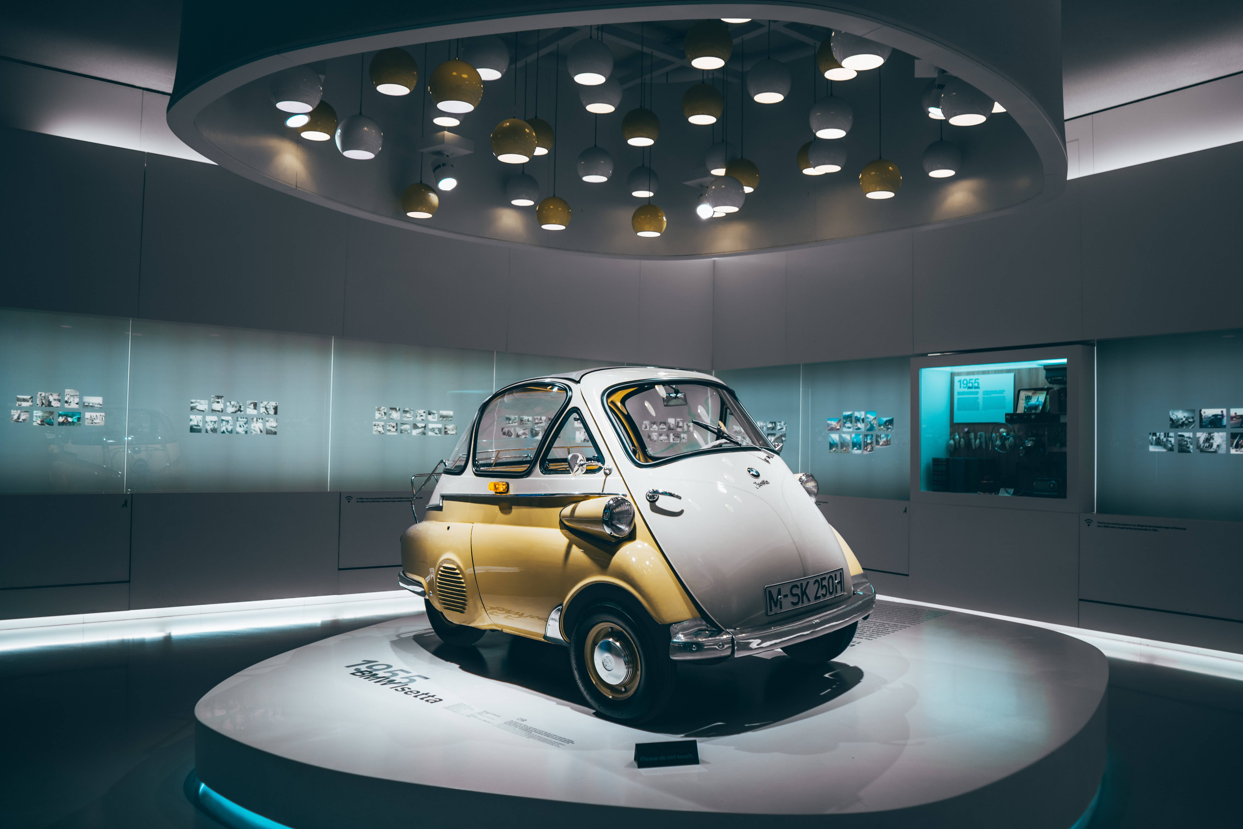 桌布 壁紙 — 一輛復古金色和白色的BMW Isetta微型車在現代懸掛燈光下的圓形平台上展示，位於時尚的博物館展覽中。