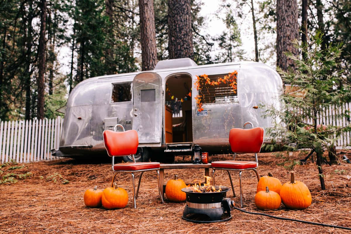 Fall Camping 桌布 壁紙 — 一輛復古銀色Airstream拖車停在松樹林中，橙色椅子和南瓜圍繞著營火。