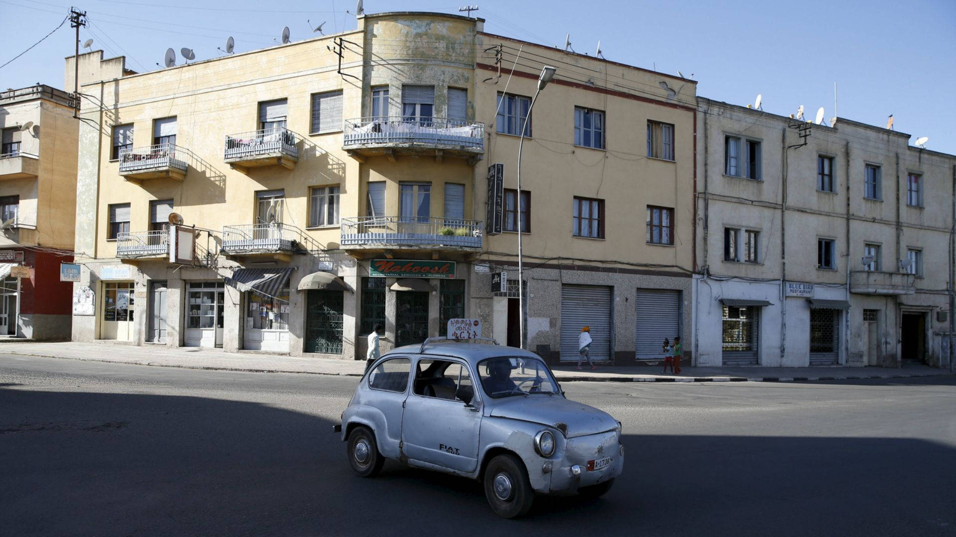 Eritrea 桌布 壁紙 — 一輛復古銀色飛雅特500在陽光明媚的街道上行駛，街道兩旁是帶有陽台和衛星天線的風化黃色建築。