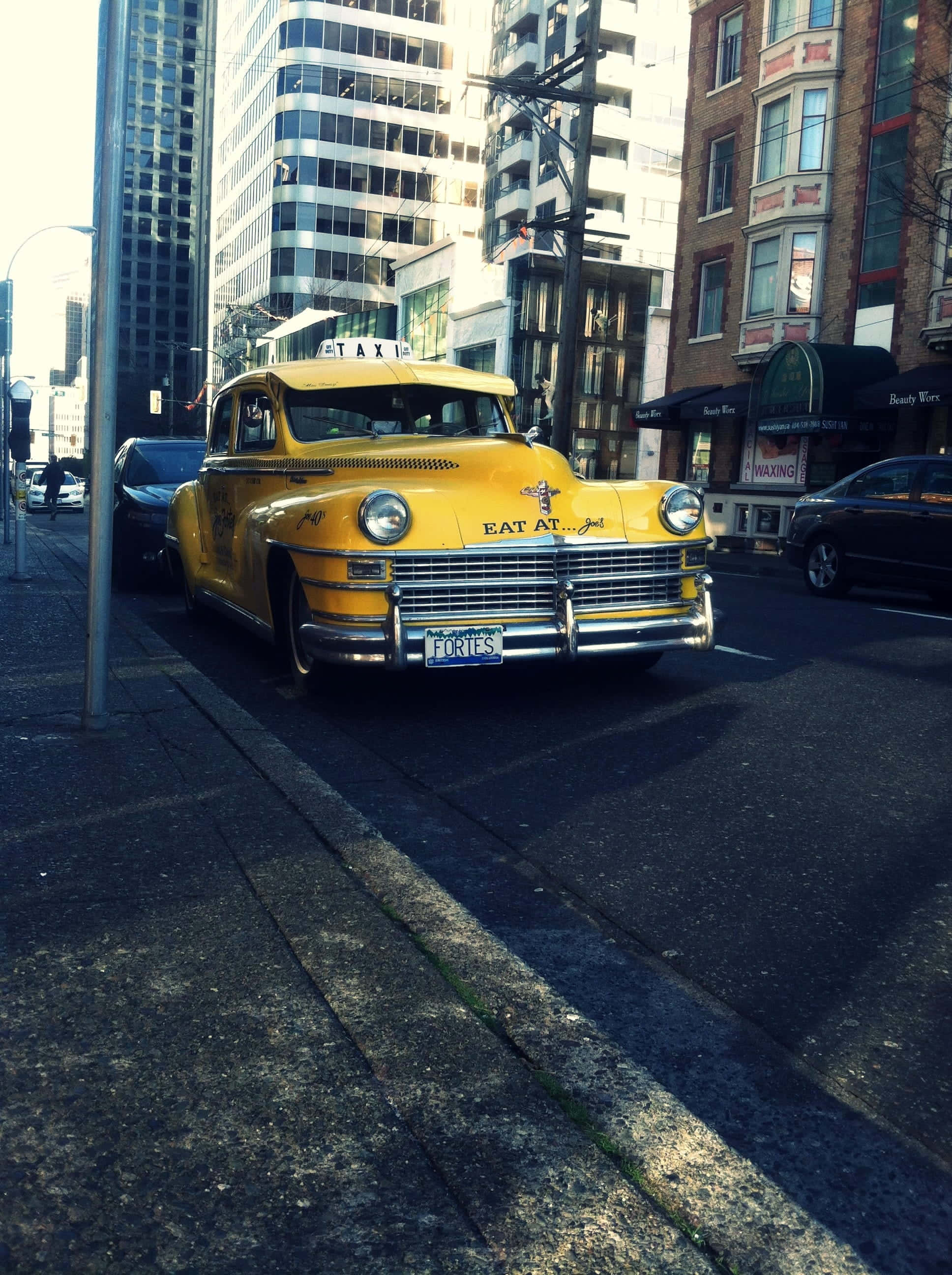 Yellow Cab 桌布 壁紙 — 一輛復古黃色計程車，水箱護罩上寫著「EAT AT...」，停放在陽光普照的街道上，兩旁是現代建築和磚造建築。