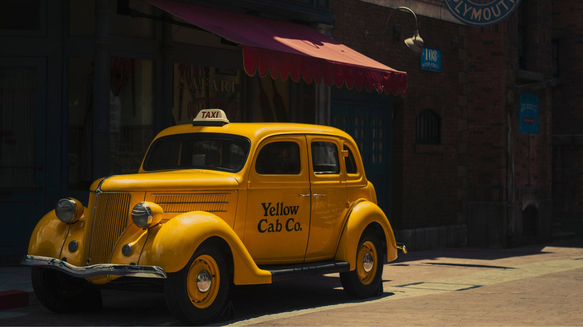 taxi 桌布 壁紙 — 一輛復古黃色計程車停泊在陽光明媚的鵝卵石街道上，旁邊是一棟帶紅色遮陽篷的磚砌建築。