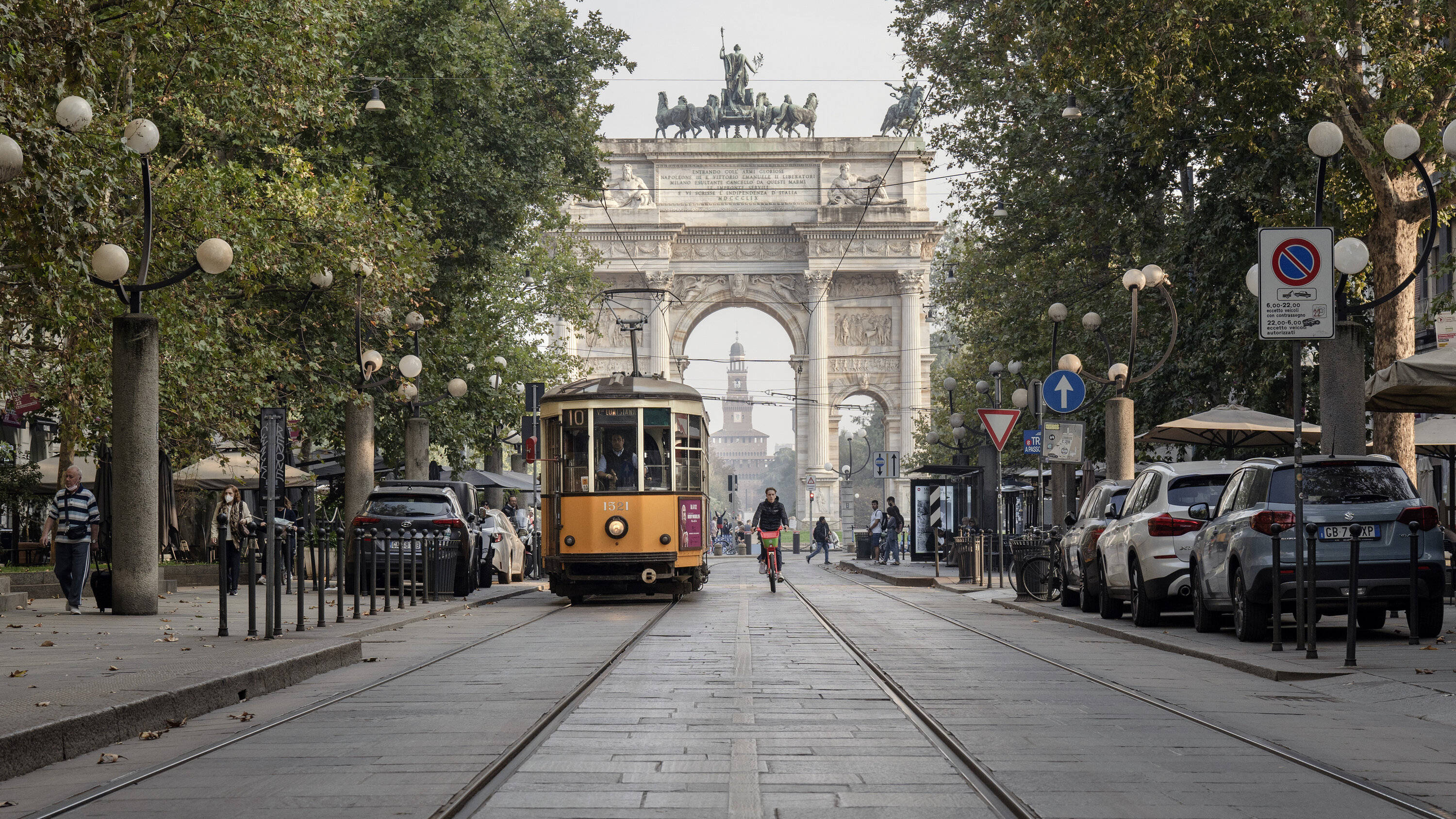 Milan 桌布 壁紙 — 一輛復古黃色電車在柔和日光下，沿著綠樹成蔭的城市街道滑行，駛向一座宏偉的石拱門。