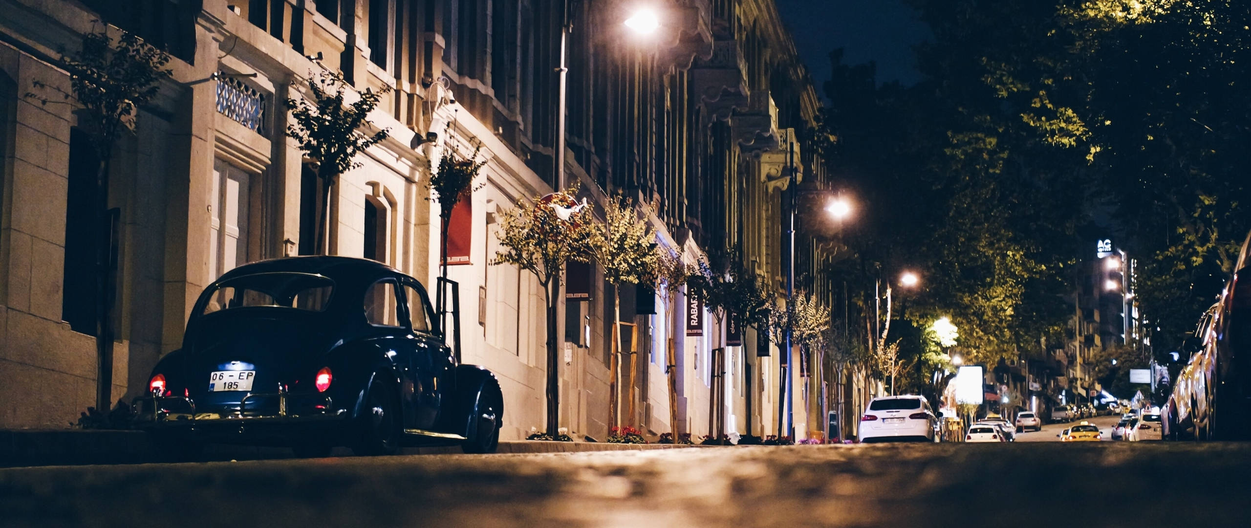 lights 桌布 壁紙 — 一輛復古黑色汽車在夜間停泊在溫暖照明的城市街道上，街道兩旁種植樹木和經典建築。