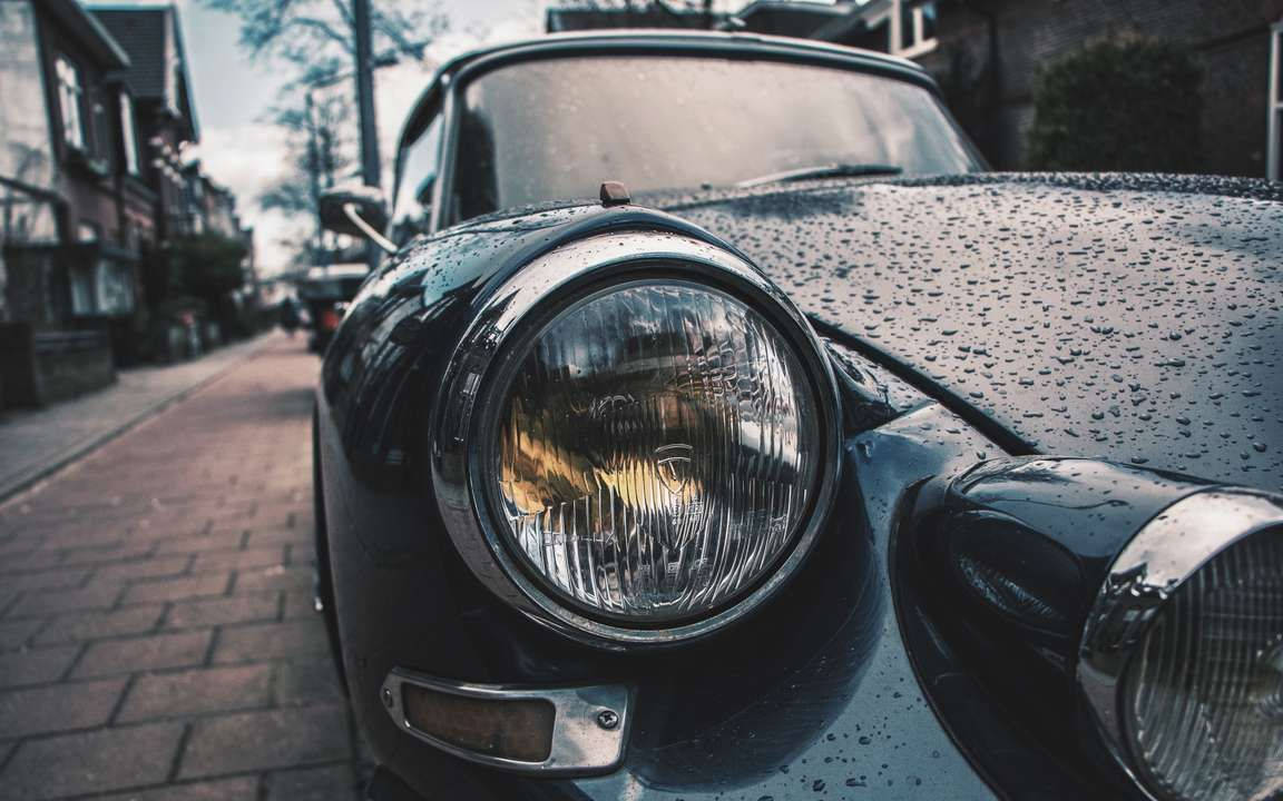高清汽車 桌布 壁紙 — 一輛復古黑色汽車的車頭燈特寫，引擎蓋上閃爍著雨滴，停泊在濕漉漉的磚砌街道上。