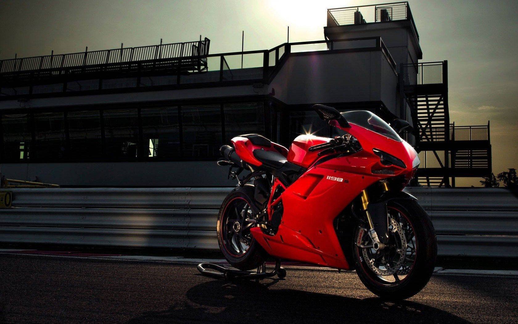 機車 桌布 壁紙 — 一輛搶眼的紅色Ducati 1198摩托車在金色夕陽下閃耀，背景是賽道上現代看台建築的剪影。