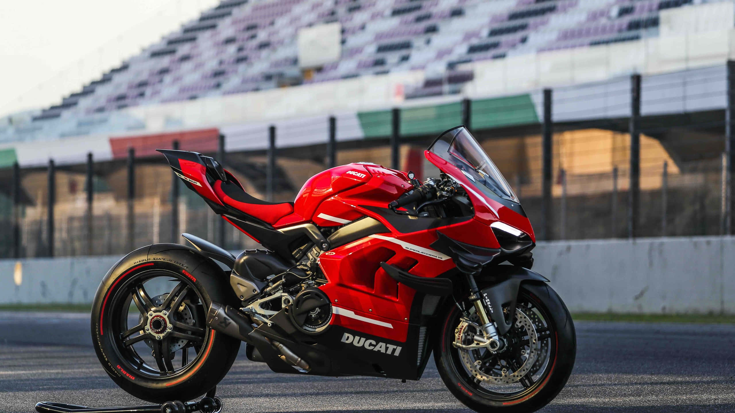 桌布 壁紙 — 一輛搶眼的紅色Ducati機車在柔和日光下，以觀眾席為背景，在空曠的賽道上閃閃發光。