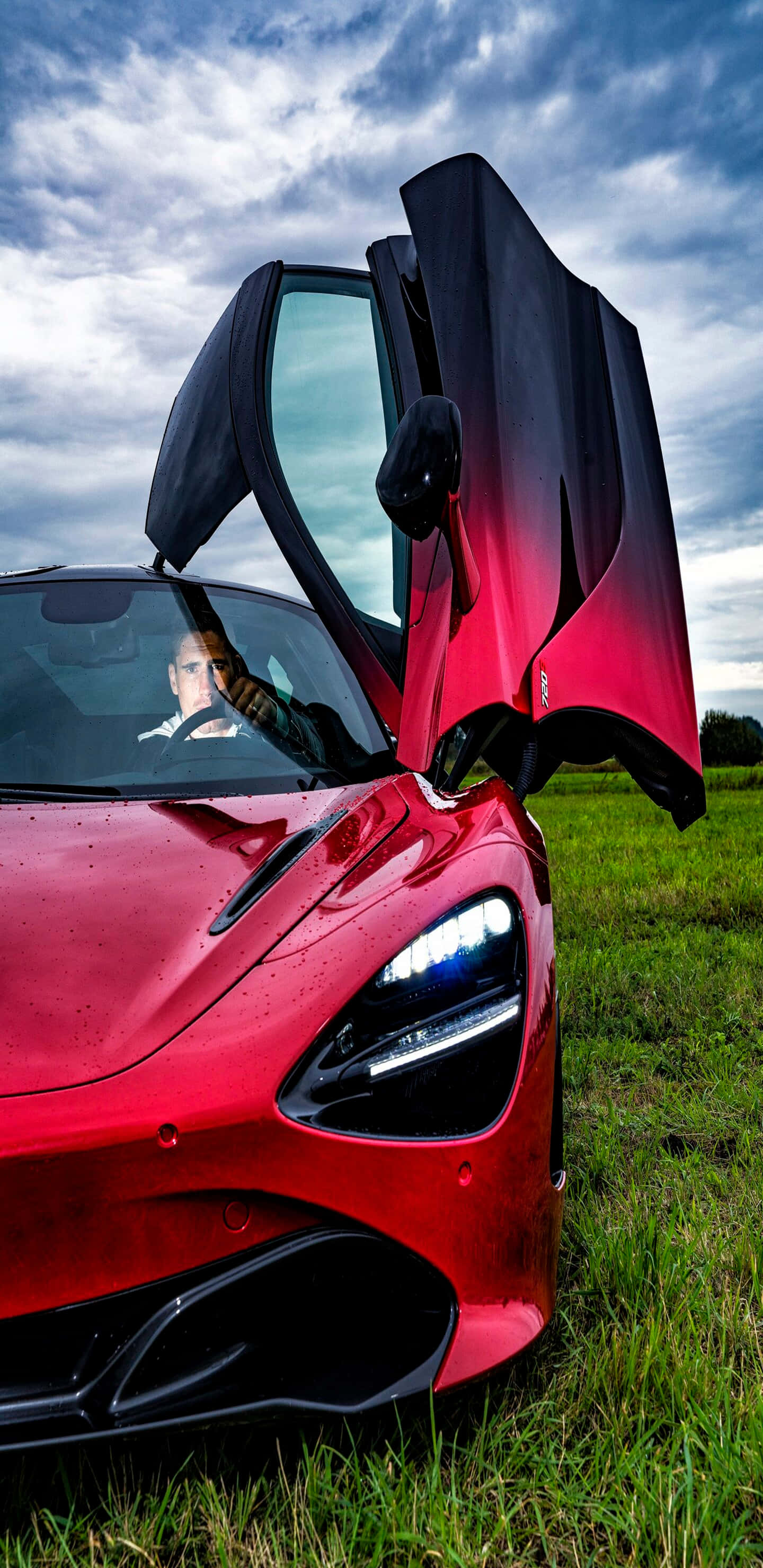 Pixel 3xl Mclaren 720s 桌布 壁紙 — 一輛搶眼的紅色McLaren跑車，鷗翼車門打開，停在草地田野上，天空多雲。