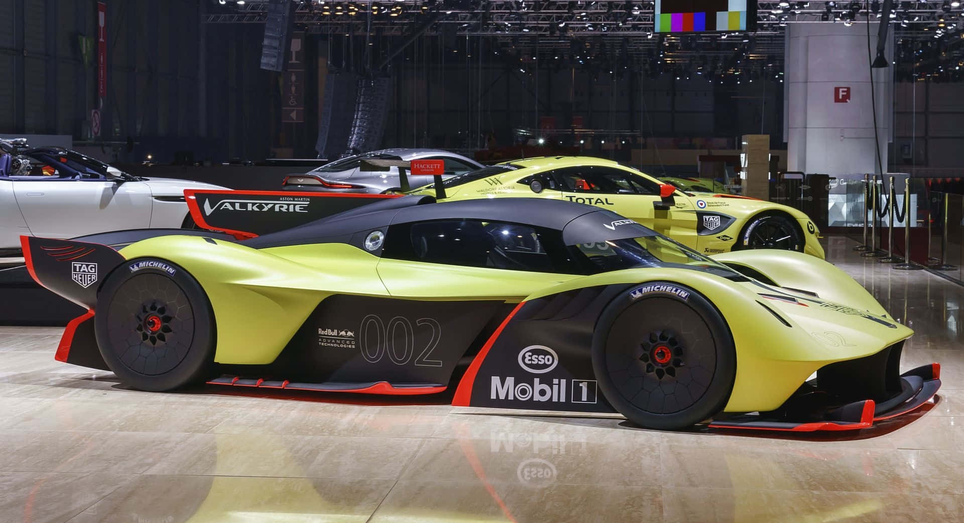 Aston Martin Valkyrie 桌布 壁紙 — 一輛搶眼的萊姆綠色Aston Martin Valkyrie賽車，帶有黑色和紅色點綴，在車展上展出，背景是其他高性能車輛。
