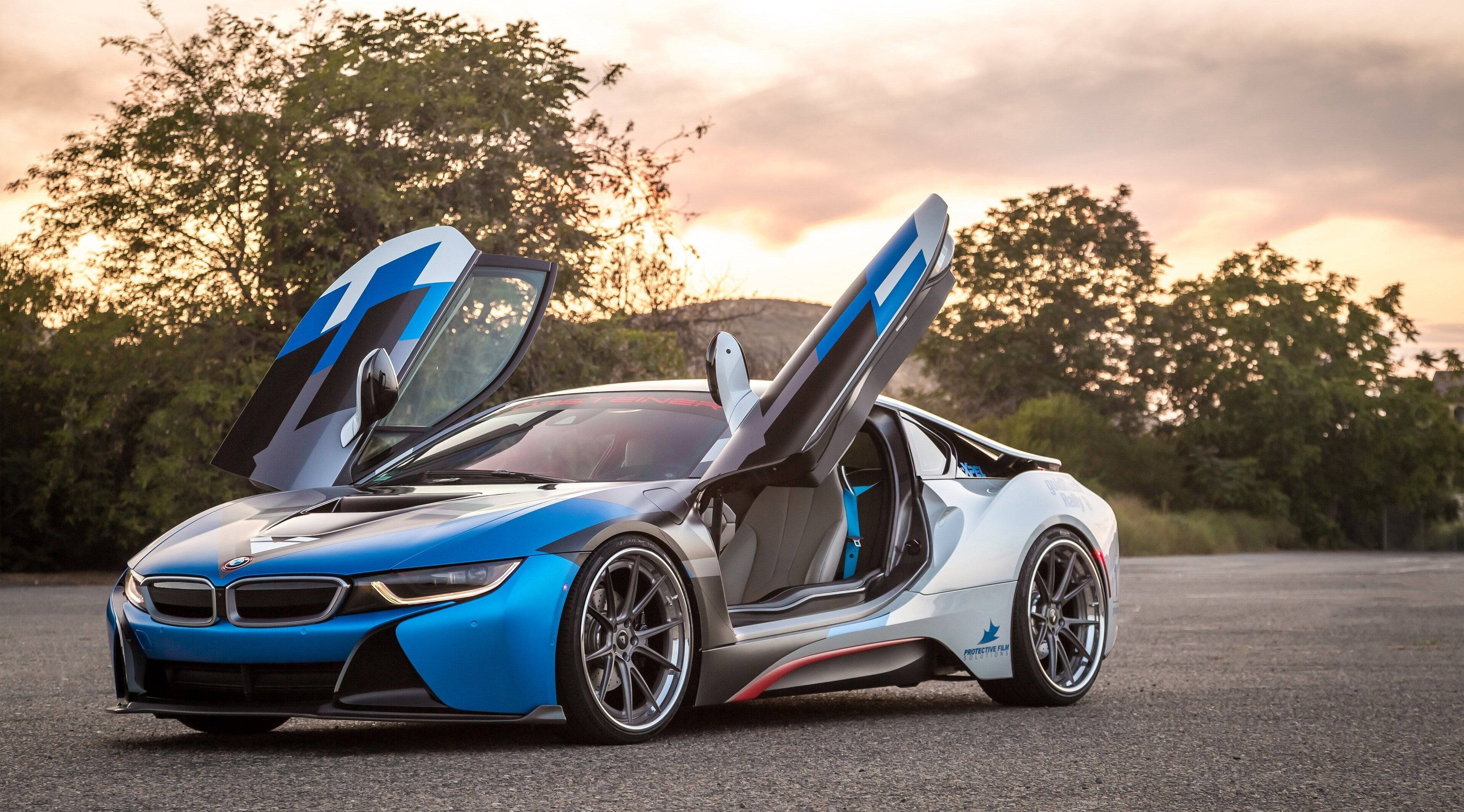 4K BMW 壁紙 桌布 壁紙 — 一輛搶眼的藍白色寶馬i8跑車，鷗翼車門打開，停在夕陽下的瀝青路上，背景有樹木。