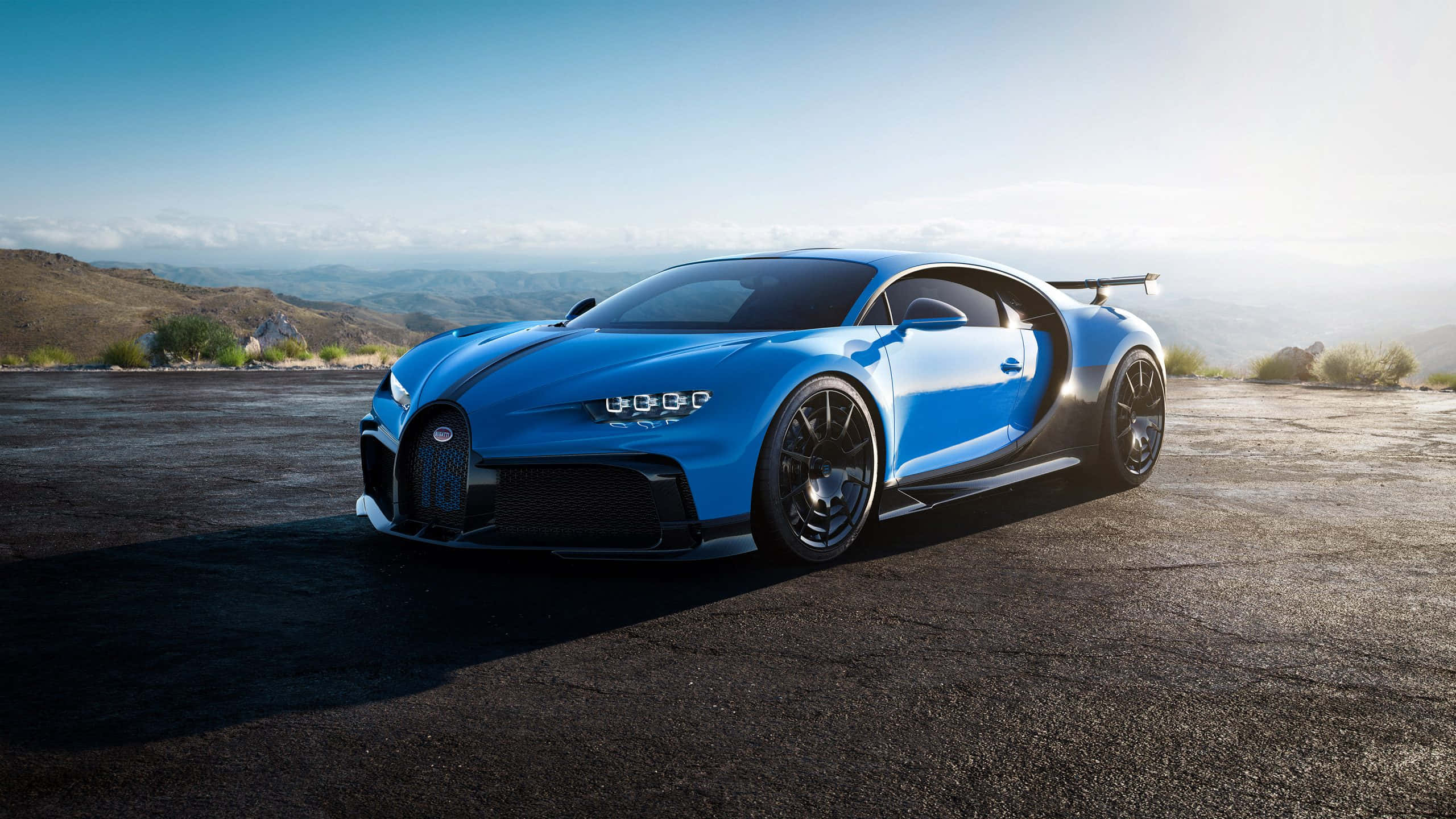 Best Bugatti 桌布 壁紙 — 一輛搶眼的藍色Bugatti Chiron Super Sport 300+停泊在陽光明媚的瀝青觀景點，遠山在晴朗的天空下清晰可見。