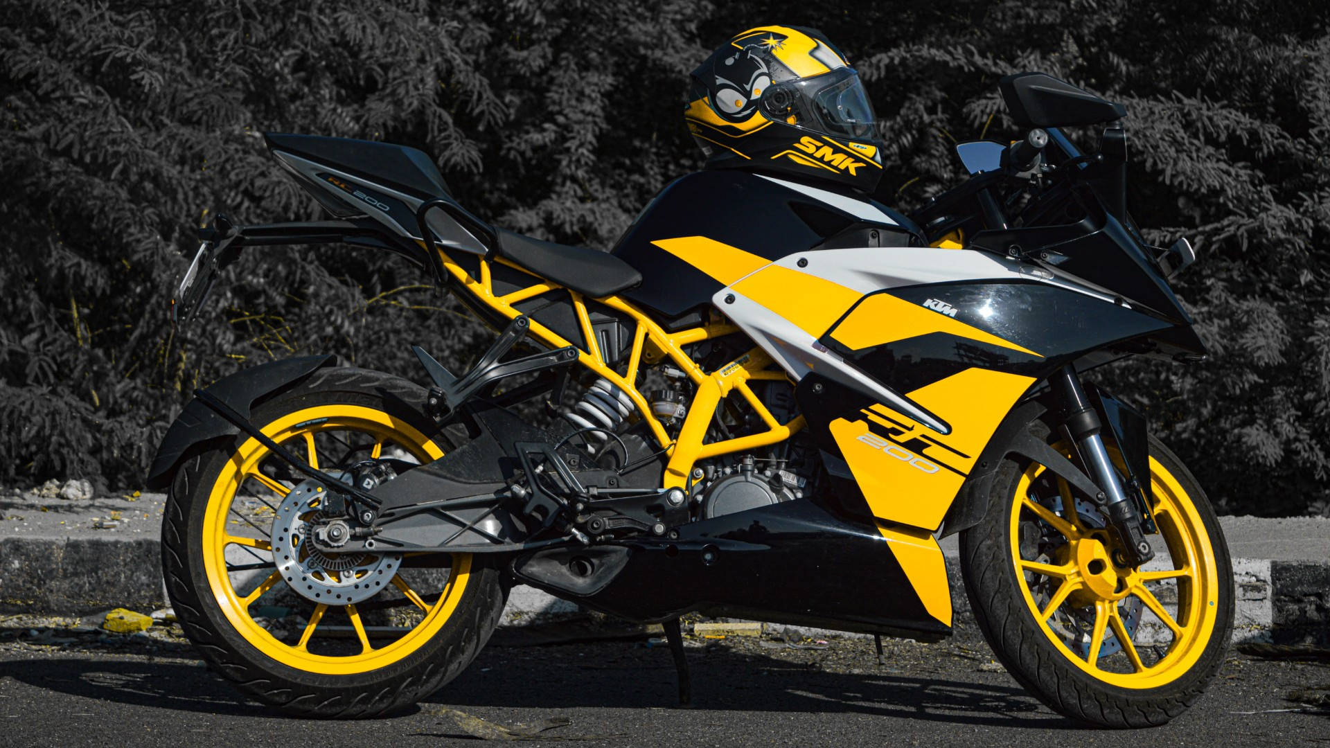 KTM RC 200 桌布 壁紙 — 一輛搶眼的黃黑配色運動機車，連同一個相配的安全帽，停放在鋪砌道路上，旁邊有深色常綠樹。