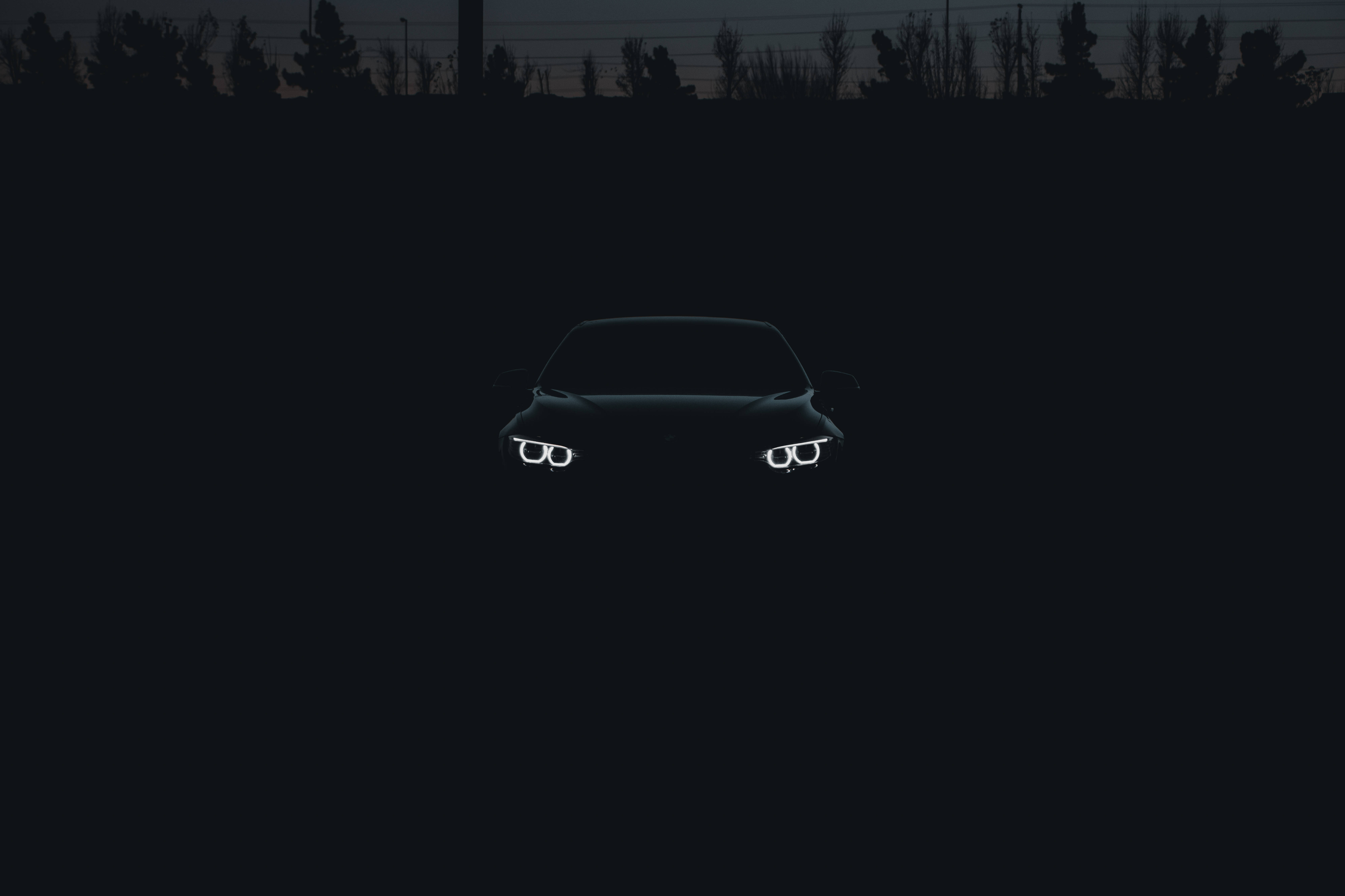 Black Bmw 桌布 壁紙 — 一輛時尚汽車的發光車頭燈在黃昏時分，以剪影樹林為背景，刺破黑暗駛近。