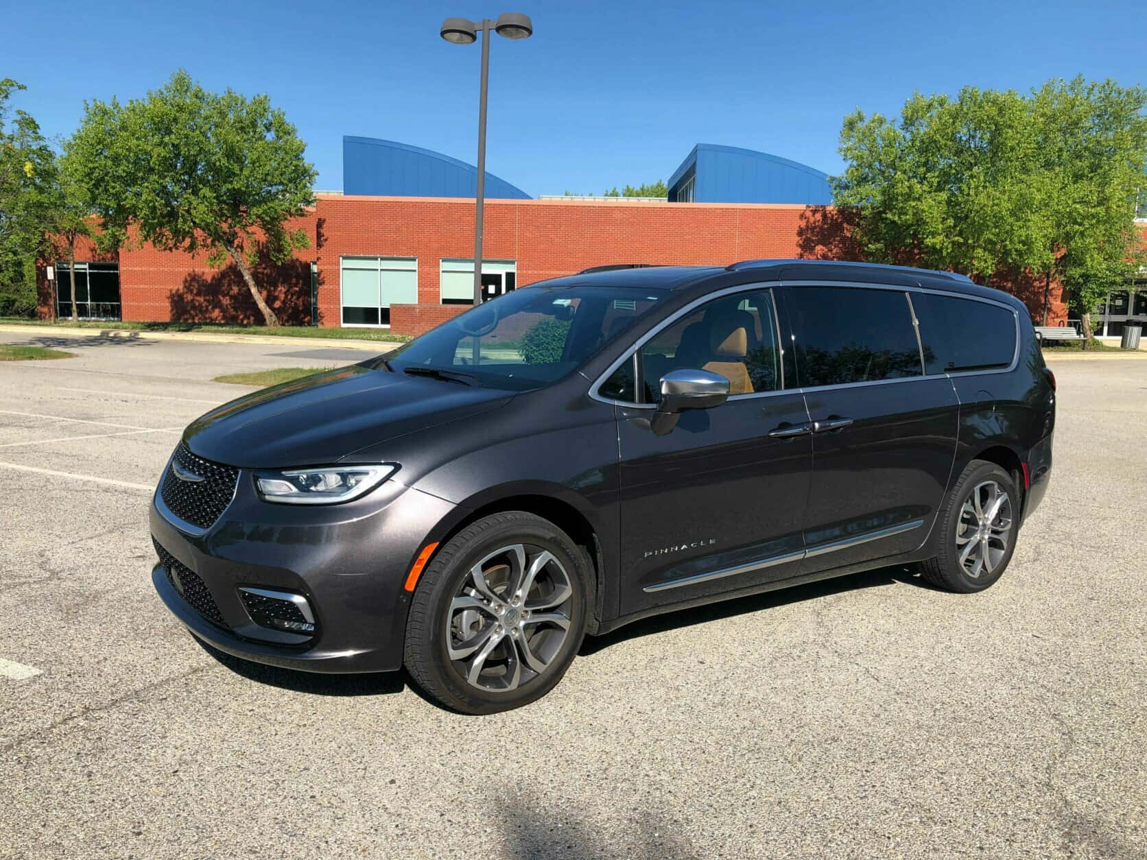 Chrysler Pacifica 桌布 壁紙 — 一輛時尚深灰色 Chrysler Pacifica 小型貨車停放在陽光普照的瀝青停車場，背景有磚造建築和綠樹。