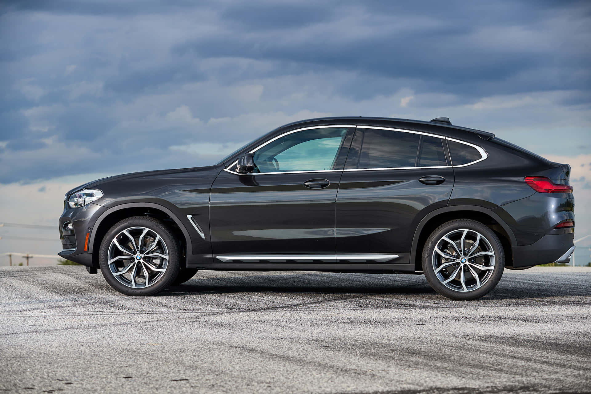 Bmw X4 桌布 壁紙 — 一輛時尚深灰色BMW X4 SUV停泊在碎石路面上，背景是戲劇性的多雲天空。