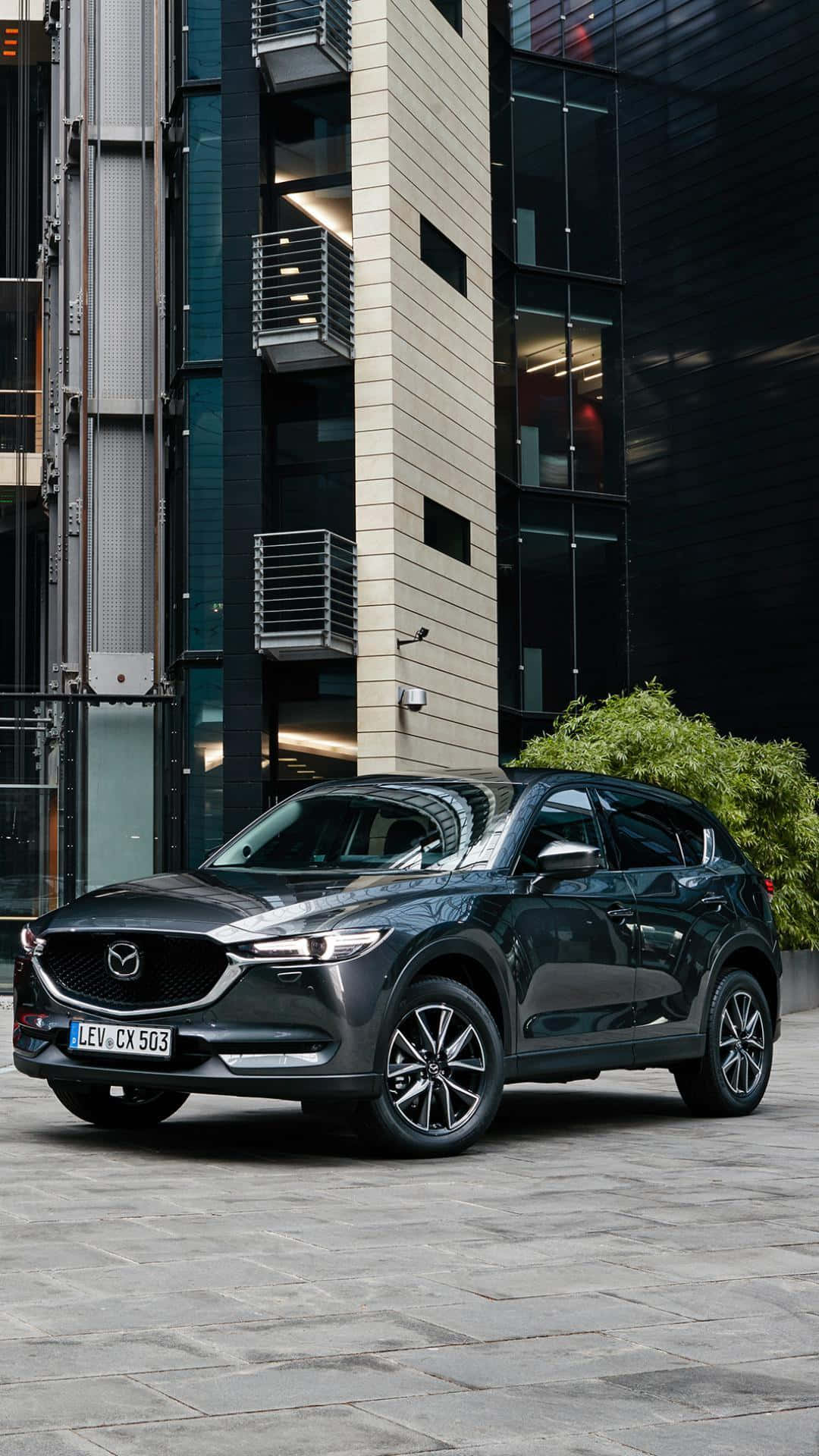 Mazda Cx-5 桌布 壁紙 — 一輛時尚深灰色Mazda CX-5 SUV停放在石頭廣場上，旁邊是一棟有綠化的現代玻璃和混凝土建築。