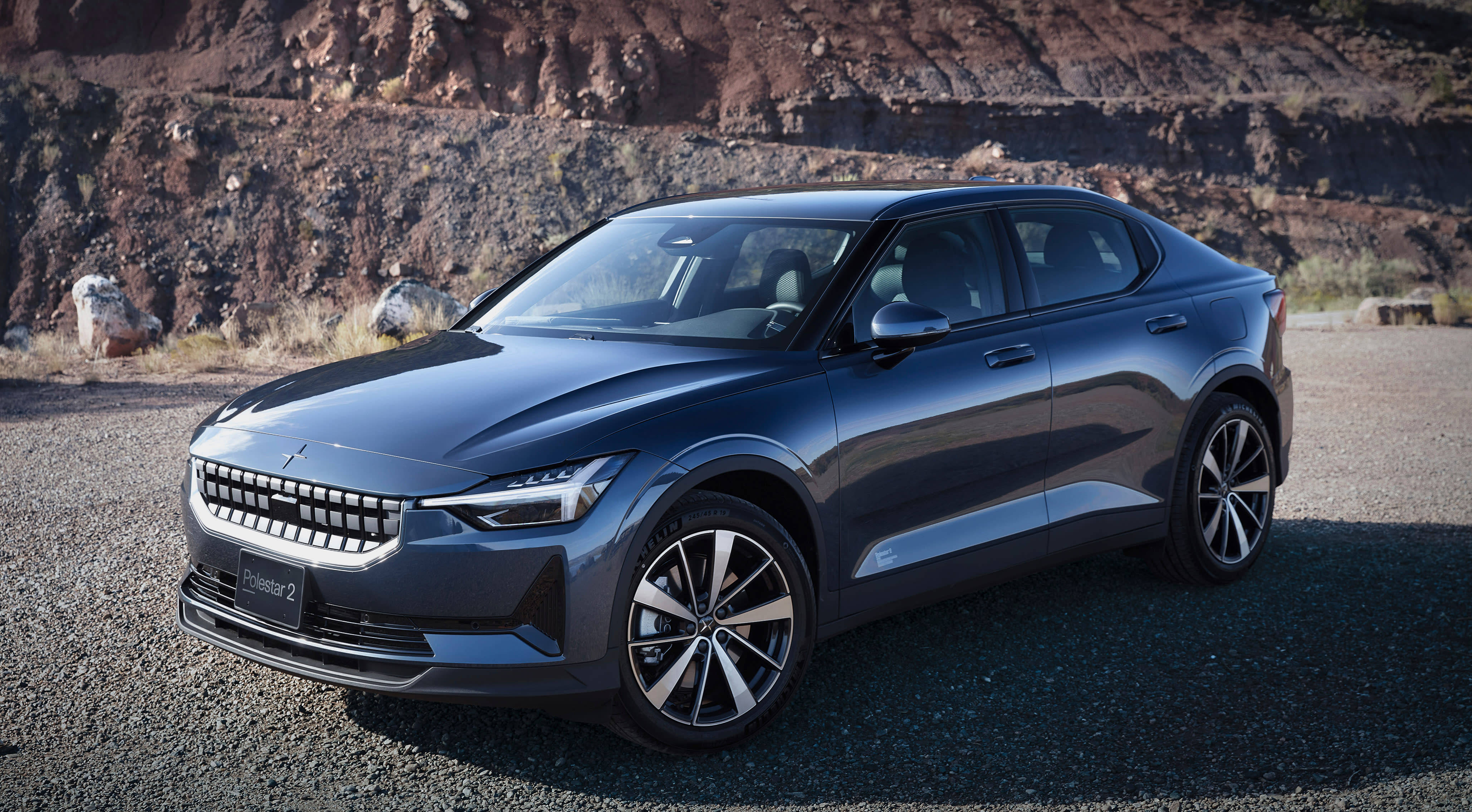 豪華車 桌布 壁紙 — 一輛時尚深灰色Polestar 2電動車停在碎石上，背景是崎嶇的紅色岩壁，在明亮的日光下。