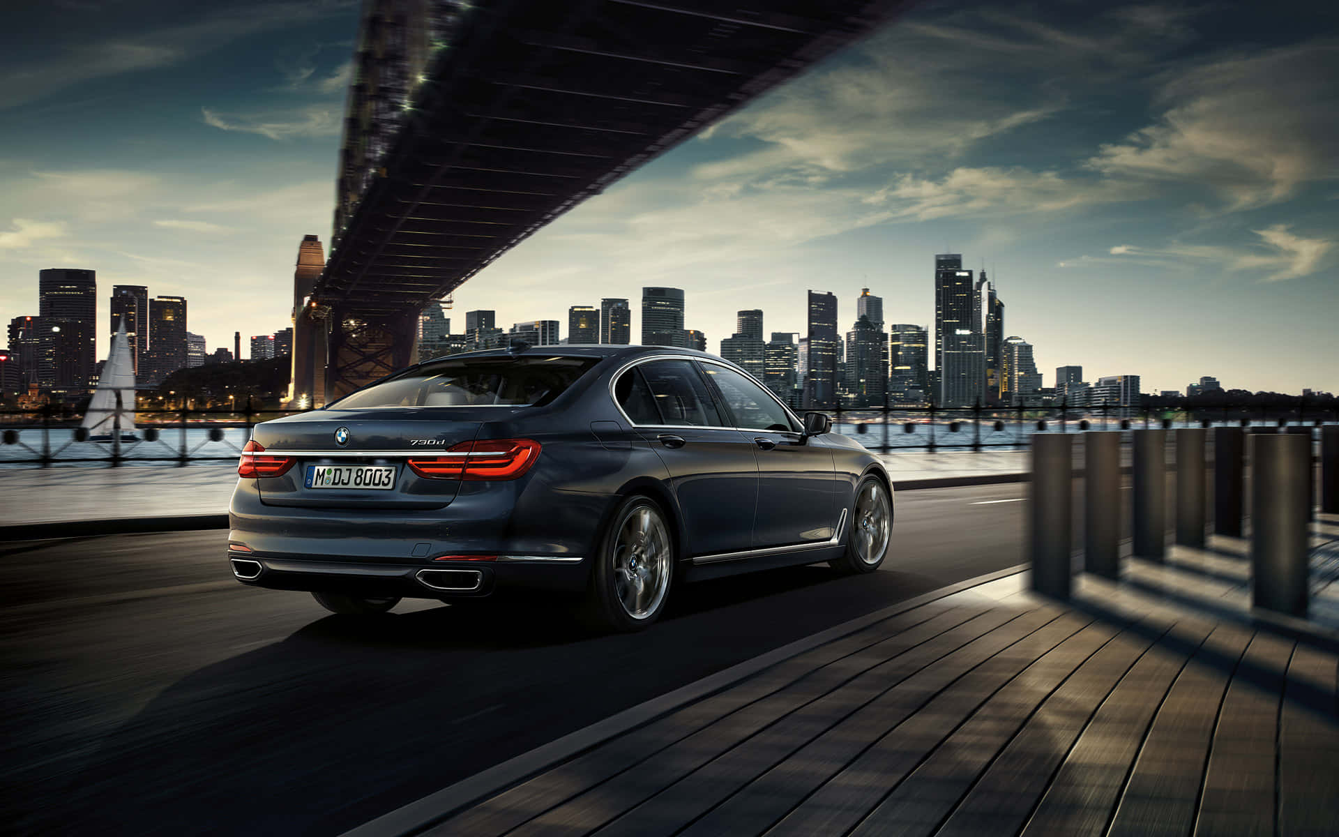 BMW 動圖 桌布 壁紙 — 一輛時尚深灰色寶馬7系列轎車在橋下行駛，現代城市天際線在黃昏時閃耀。