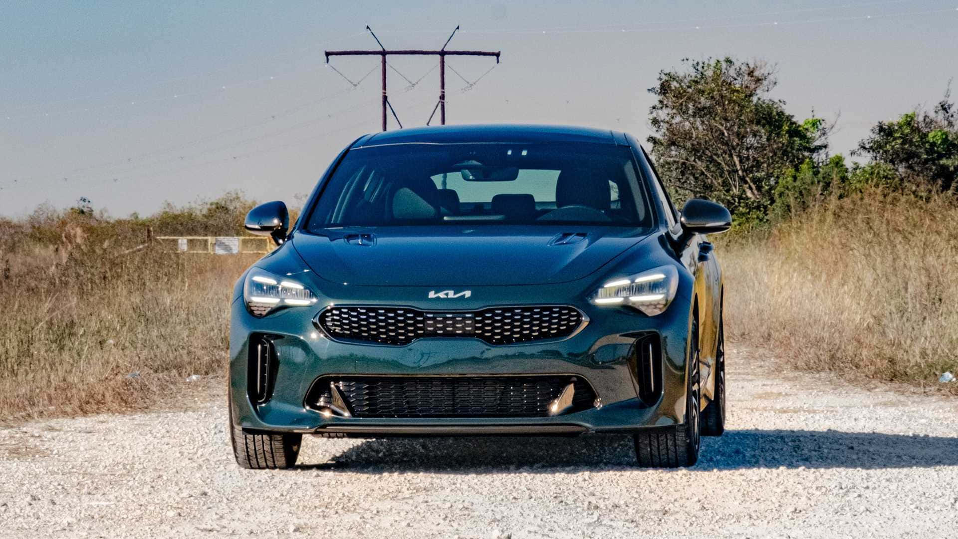 Kia Stinger 桌布 壁紙 — 一輛時尚深綠色Kia Stinger跑車在碎石路上向前，周圍有乾草和電線，天空晴朗。