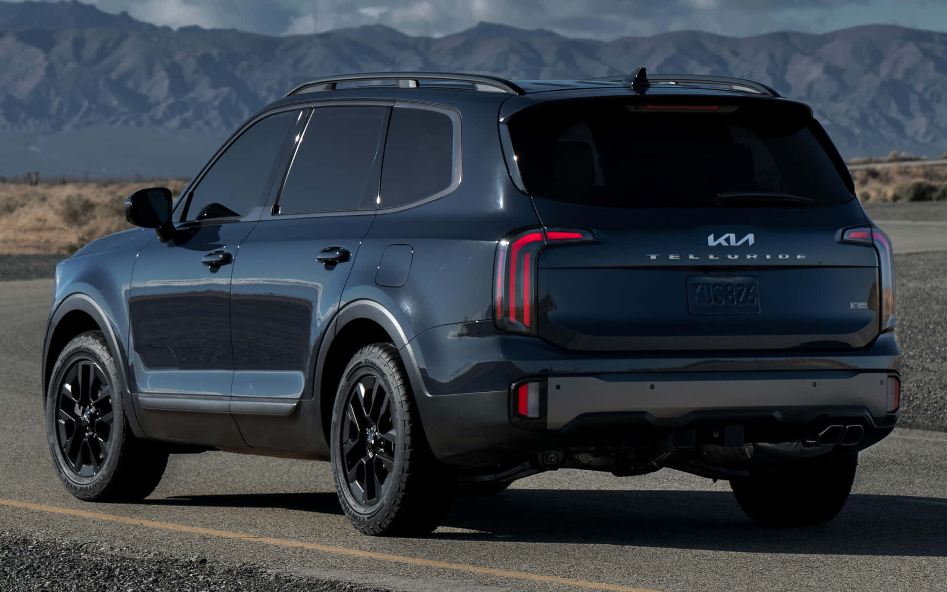 Kia Telluride 桌布 壁紙 — 一輛時尚深藍色Kia Telluride SUV停泊在沙漠公路上，背景是崎嶇的山脈，天空晴朗。