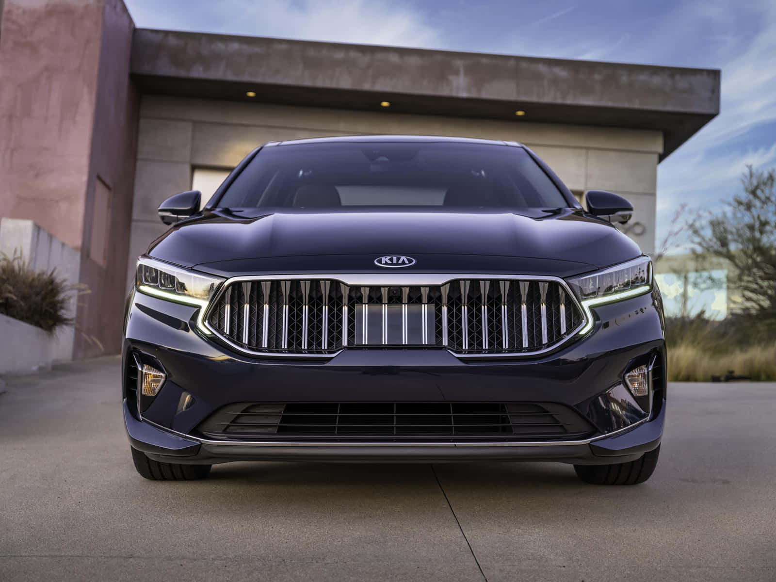 Kia Cadenza 桌布 壁紙 — 一輛時尚深藍色起亞轎車正面朝前，配有大膽格柵和鋒利頭燈，停在一棟現代混凝土建築前。