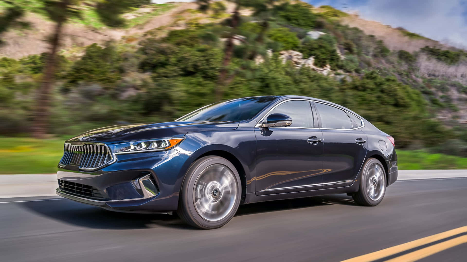 Kia Cadenza 桌布 壁紙 — 一輛時尚深藍色轎車沿著蜿蜒的山路疾馳，背景是茂密的綠樹和岩石地形，呈現動態模糊效果。