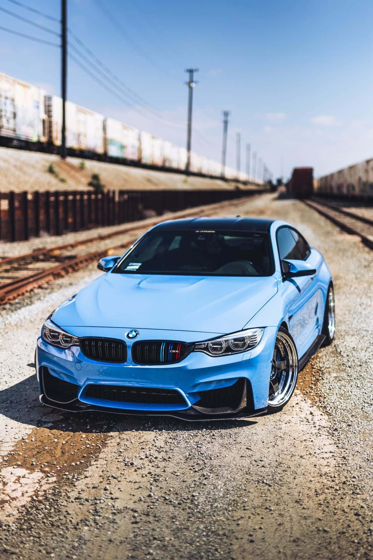 BMW 動圖 桌布 壁紙 — 一輛時尚淺藍色BMW M4雙門轎跑車停在鐵軌旁的碎石上，背景有貨運列車和晴朗的藍天。