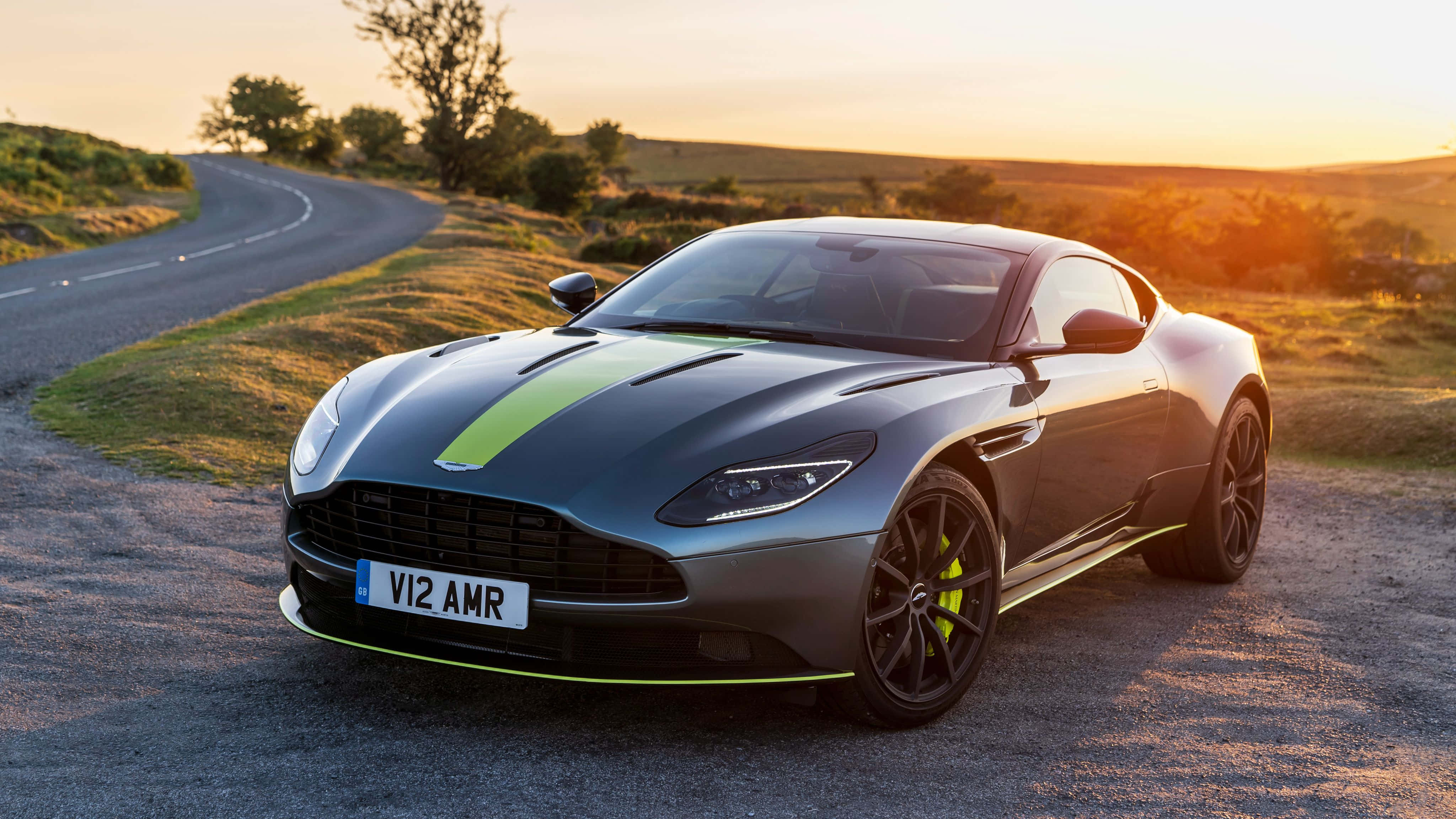 Aston Martin Db11 桌布 壁紙 — 一輛時尚灰色 Aston Martin V12 AMR 搭配萊姆綠色點綴，停放在金色日落時分的蜿蜒鄉村道路上。
