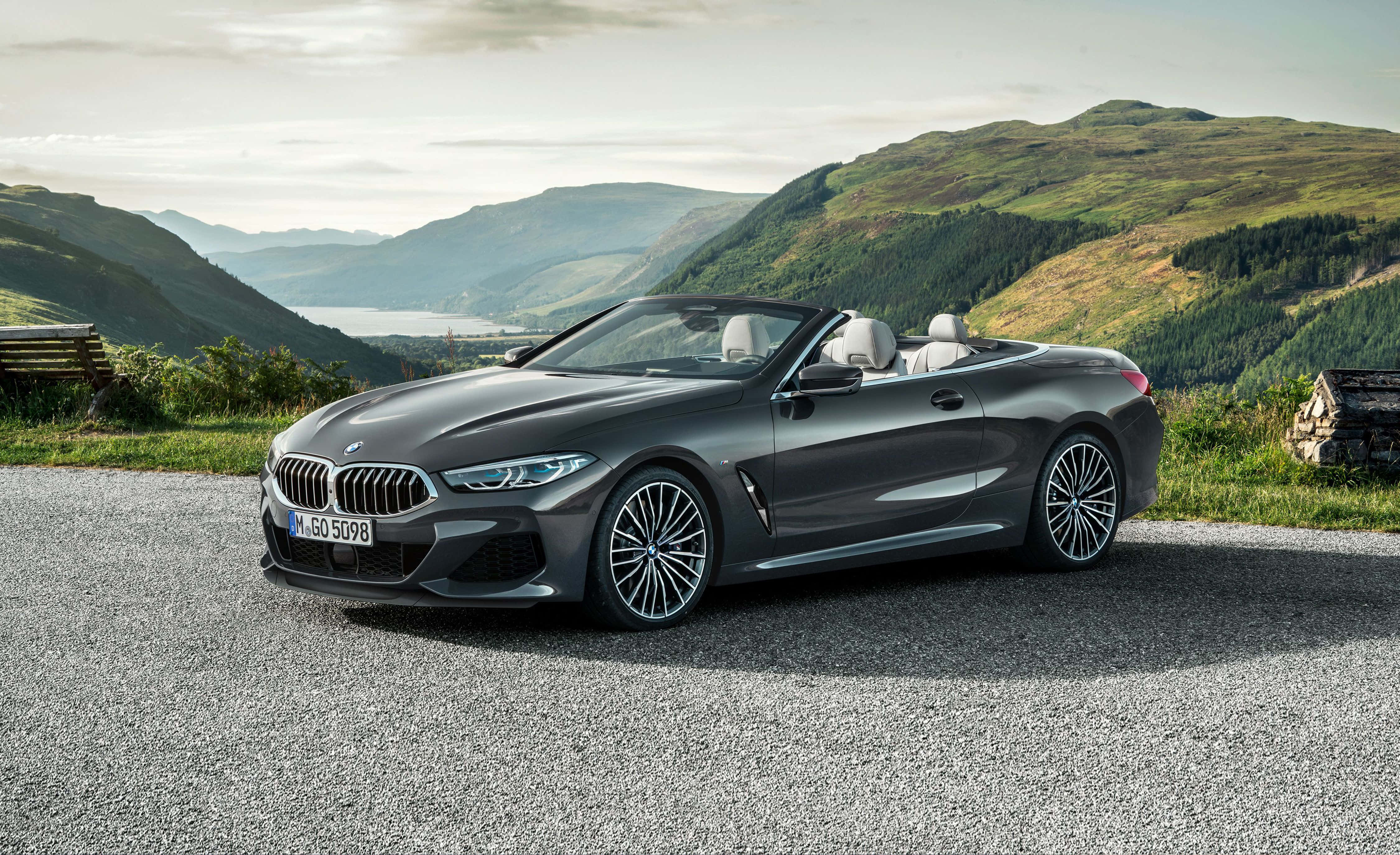 Bmw 8 Series 桌布 壁紙 — 一輛時尚灰色BMW敞篷車停泊在碎石路上，周圍是起伏的綠色山丘和遠處的湖泊，天空柔和。