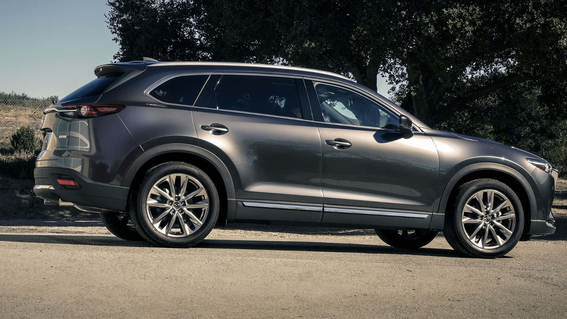 Mazda Cx-9 桌布 壁紙 — 一輛時尚灰色Mazda CX-90 SUV停泊在陽光照耀的碎石路上，背景有樹木。