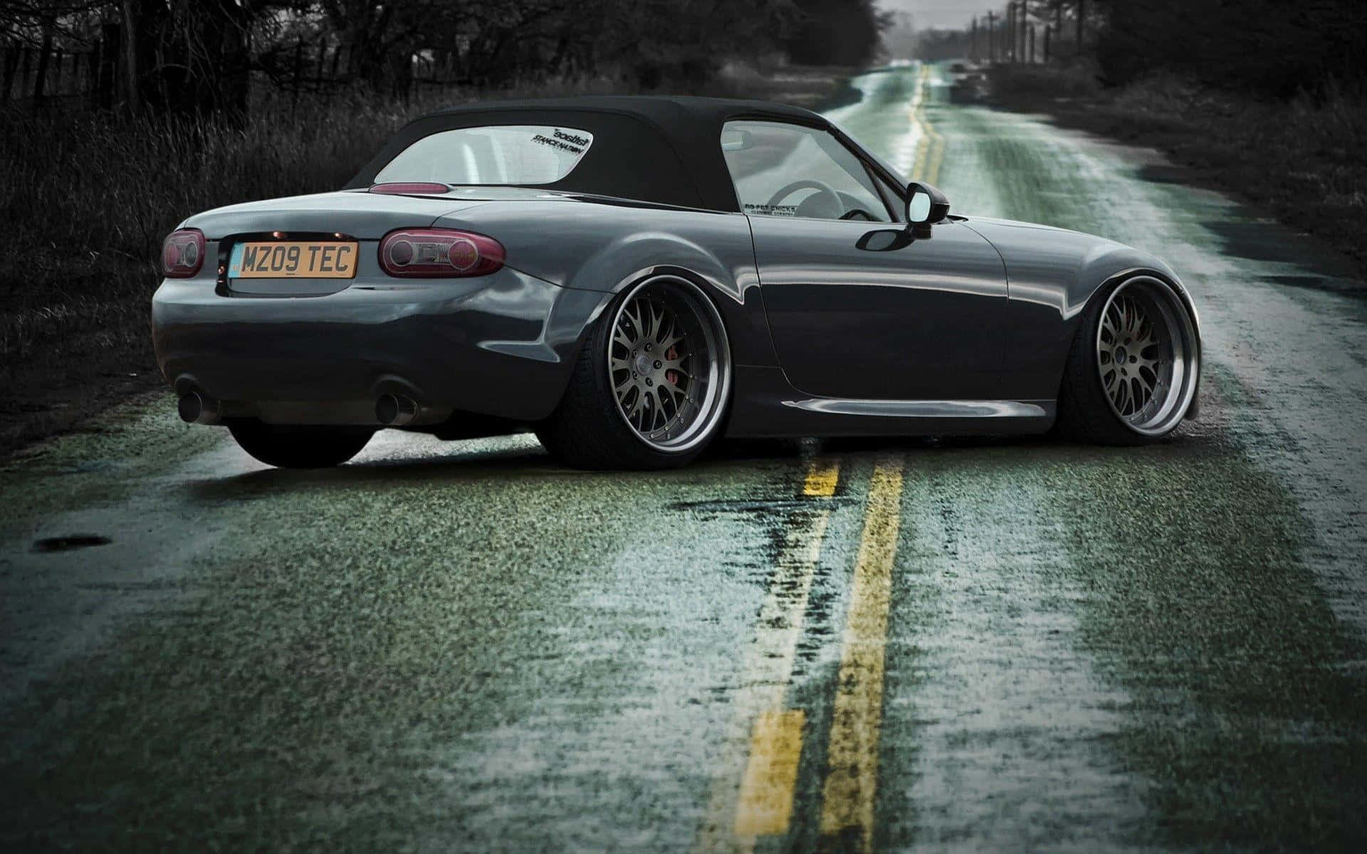 Miata 桌布 壁紙 — 一輛時尚灰色敞篷跑車配備訂製輪圈，停泊在潮濕的鄉村道路上，天空多雲。