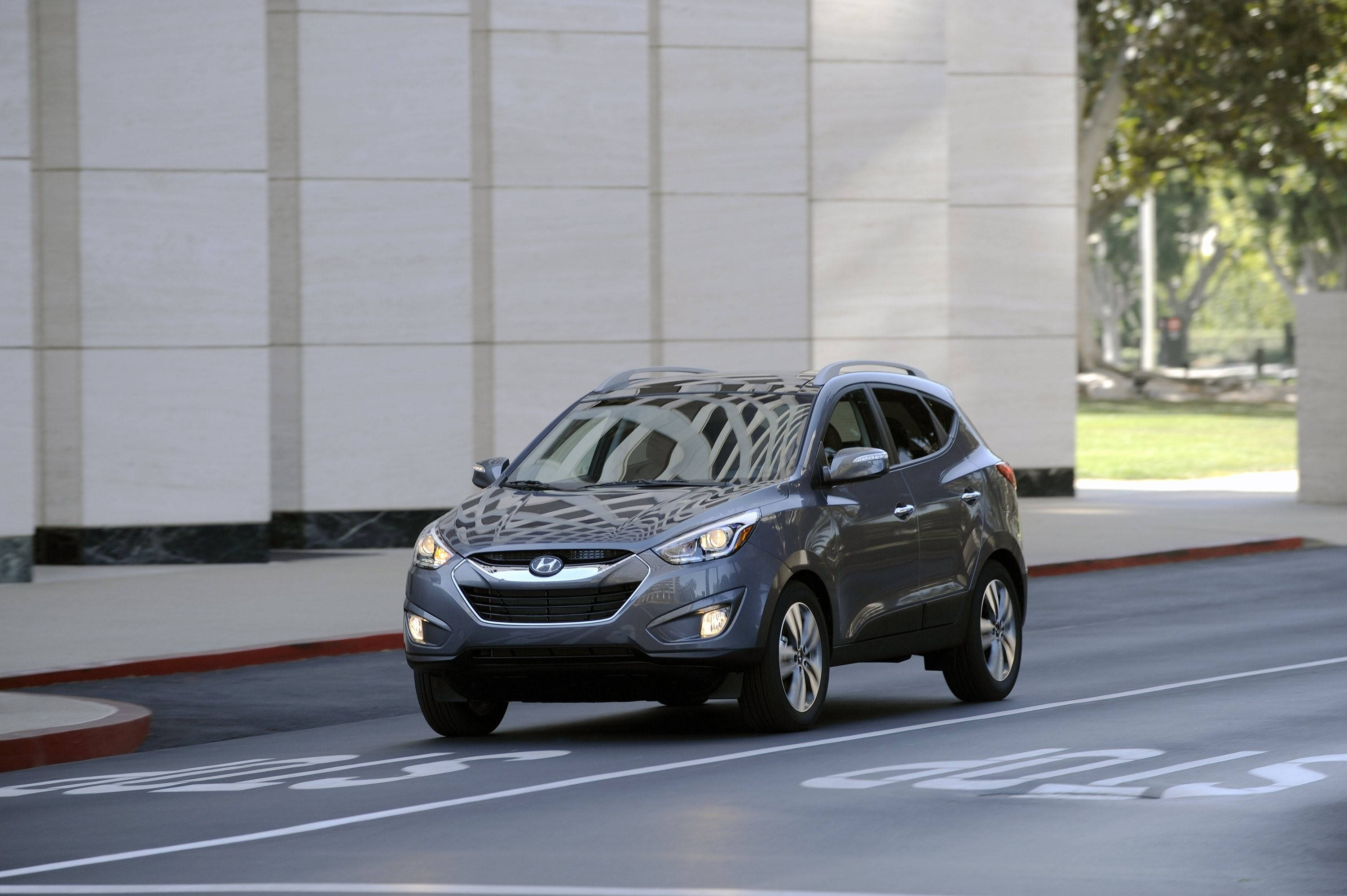 Hyundai 桌布 壁紙 — 一輛時尚灰色現代Tucson SUV在城市街道上平穩行駛，旁邊是現代石建築，背景有樹木。