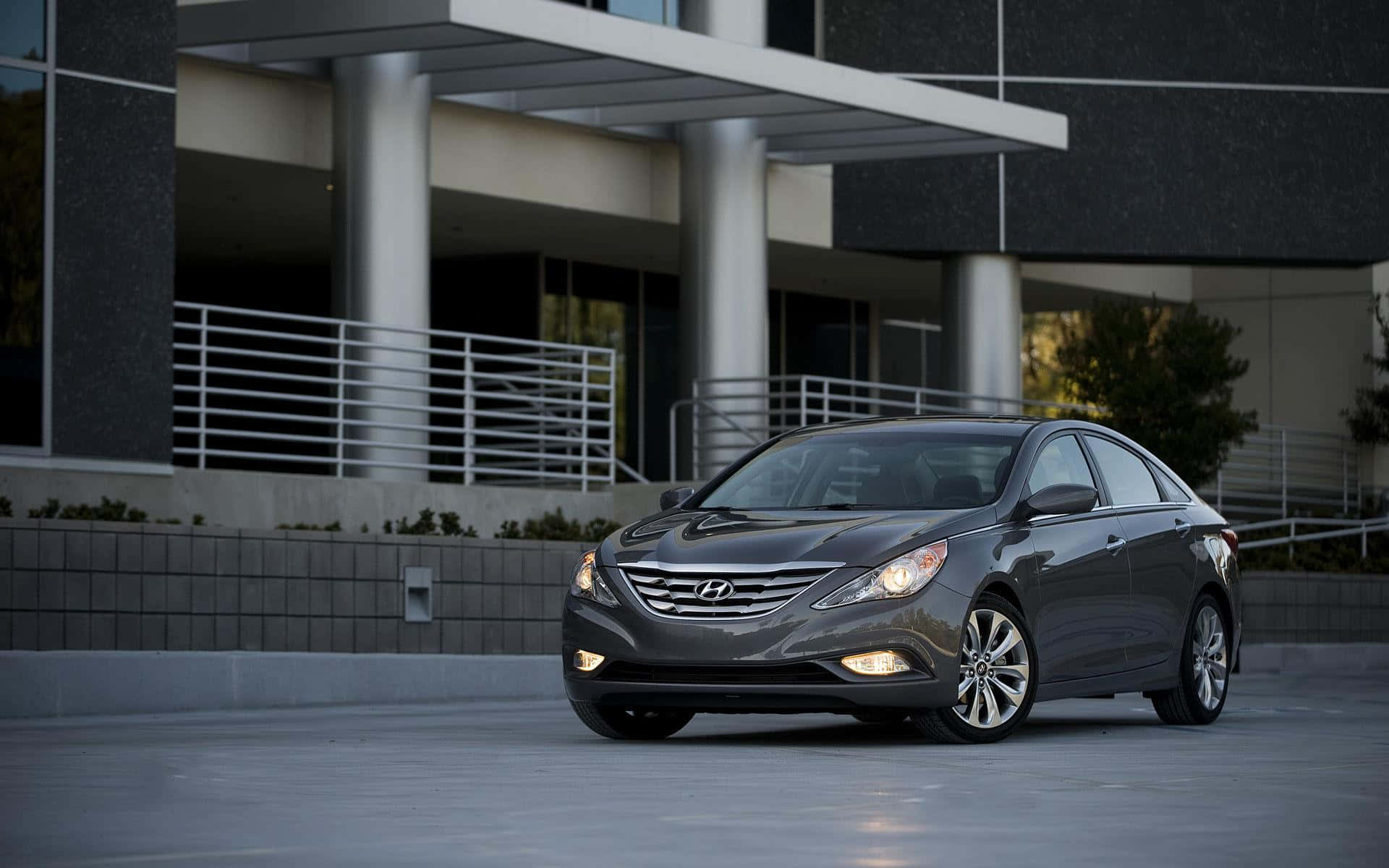 Hyundai Sonata 桌布 壁紙 — 一輛時尚灰色現代轎車停在現代建築前，車頭燈亮起。