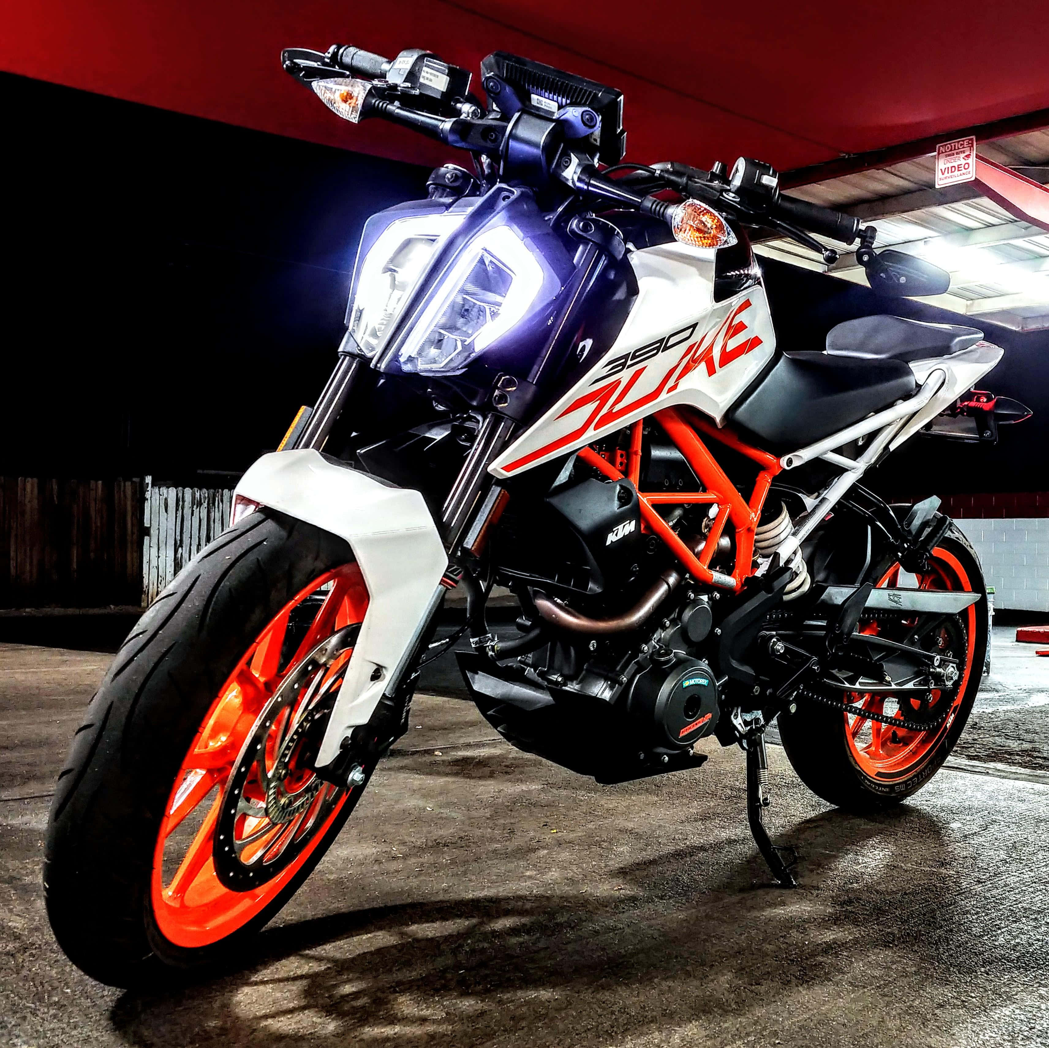 Ktm 桌布 壁紙 — 一輛時尚的KTM 390 Duke摩托車，帶有鮮豔的橙色點綴和發光的頭燈，停在昏暗車庫的紅色簷篷下。