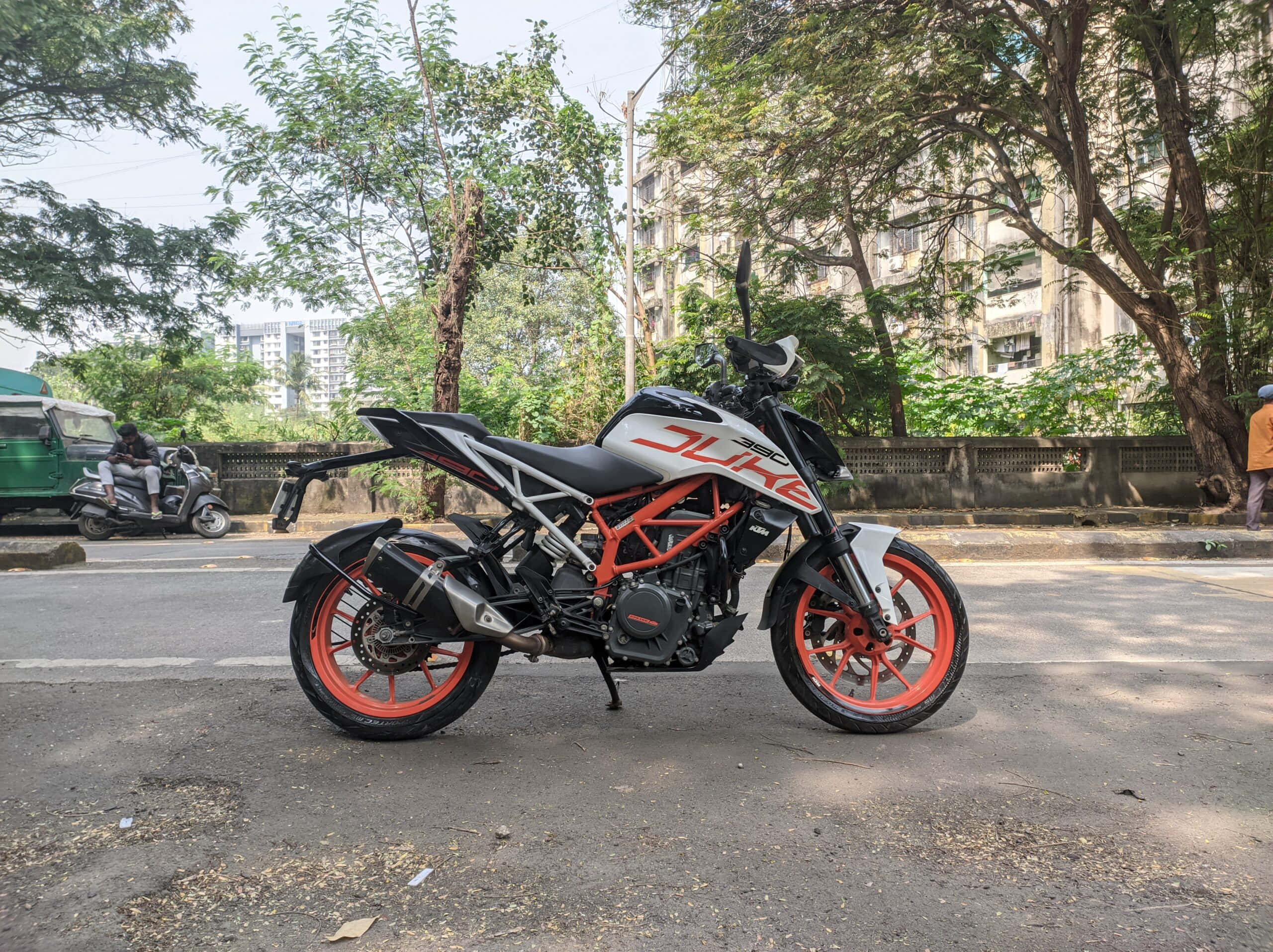 Ktm 桌布 壁紙 — 一輛時尚的KTM Duke摩托車，配備橙色車輪，停放在陽光普照的市區街道上，兩旁種滿樹木和建築物。