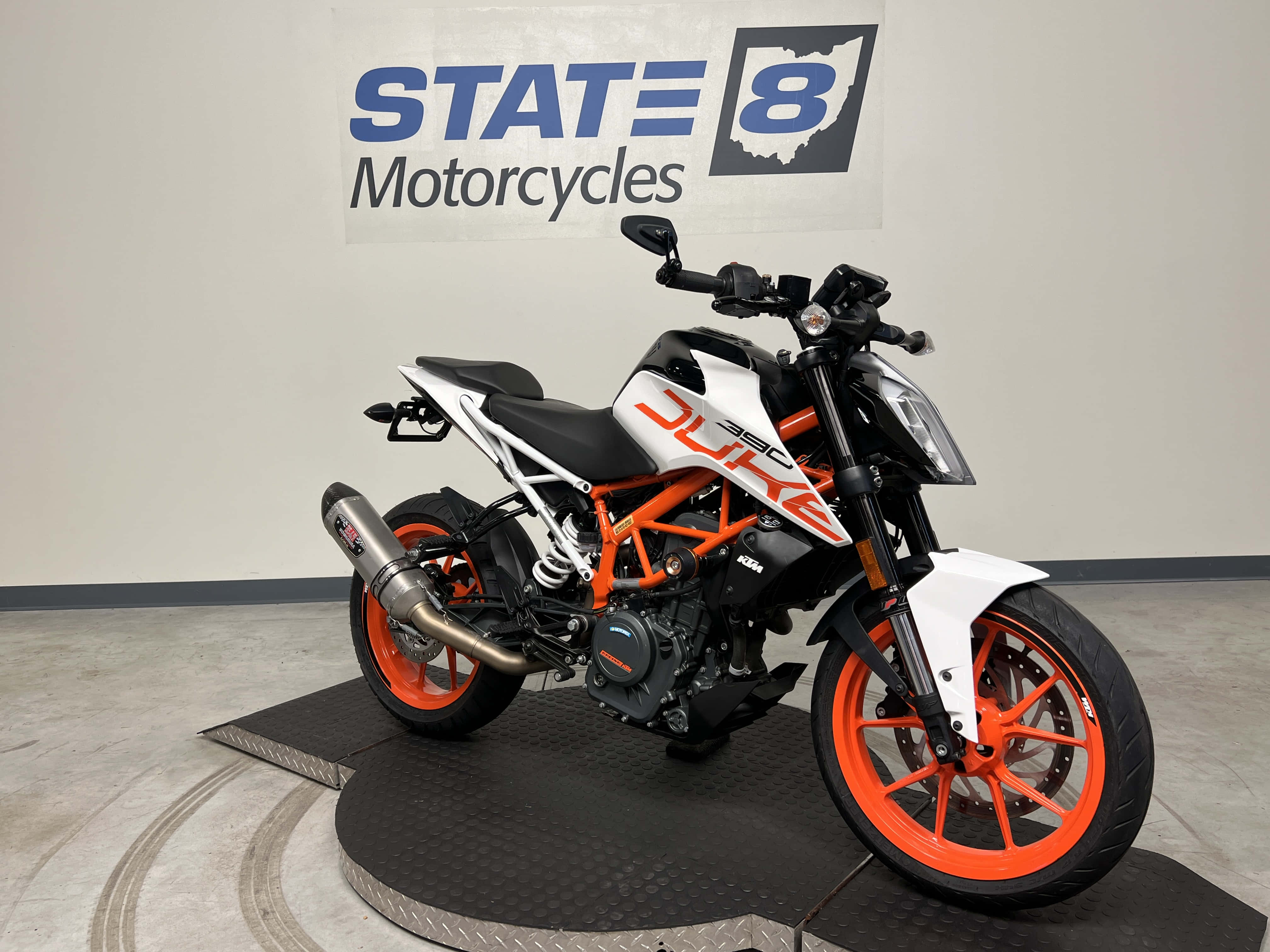 機車 桌布 壁紙 — 一輛時尚的KTM Duke機車，帶有橙色和白色點綴，停放在展廳的黑色平台上，牆上掛有State 8 Motorcycles標誌。
