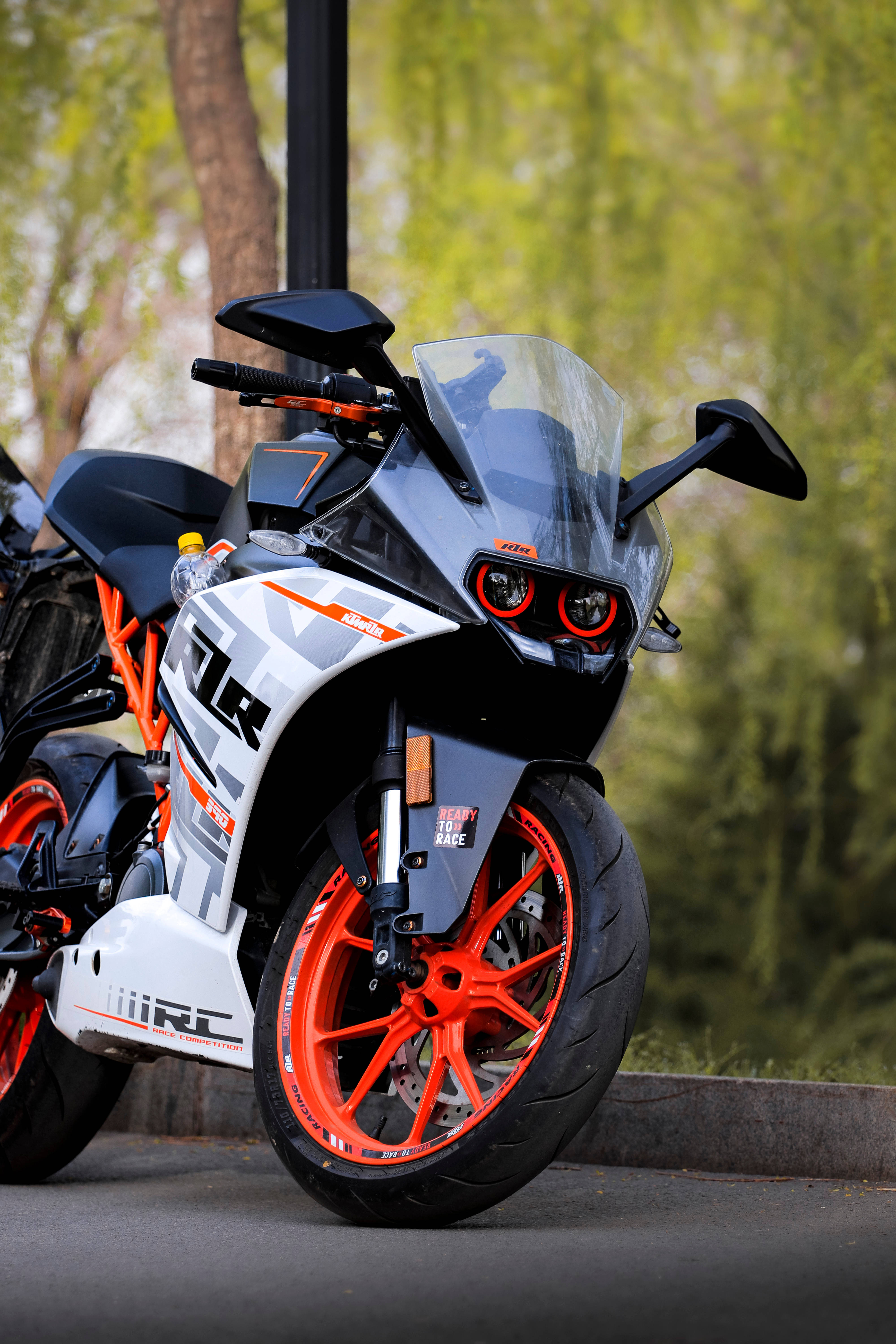 Ktm Rc 390 桌布 壁紙 — 一輛時尚的KTM RC摩托車，帶有鮮豔的橙色車輪和黑白圖案，停在鋪砌路面上，旁邊有綠樹。