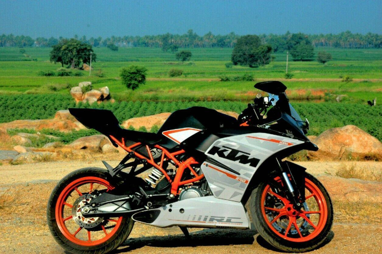 KTM 摩托車 桌布 壁紙 — 一輛時尚的KTM機車，配備橙色輪圈和銀色車架，停在翠綠田野和散落岩石間的泥土路上，背景是晴朗的藍天。