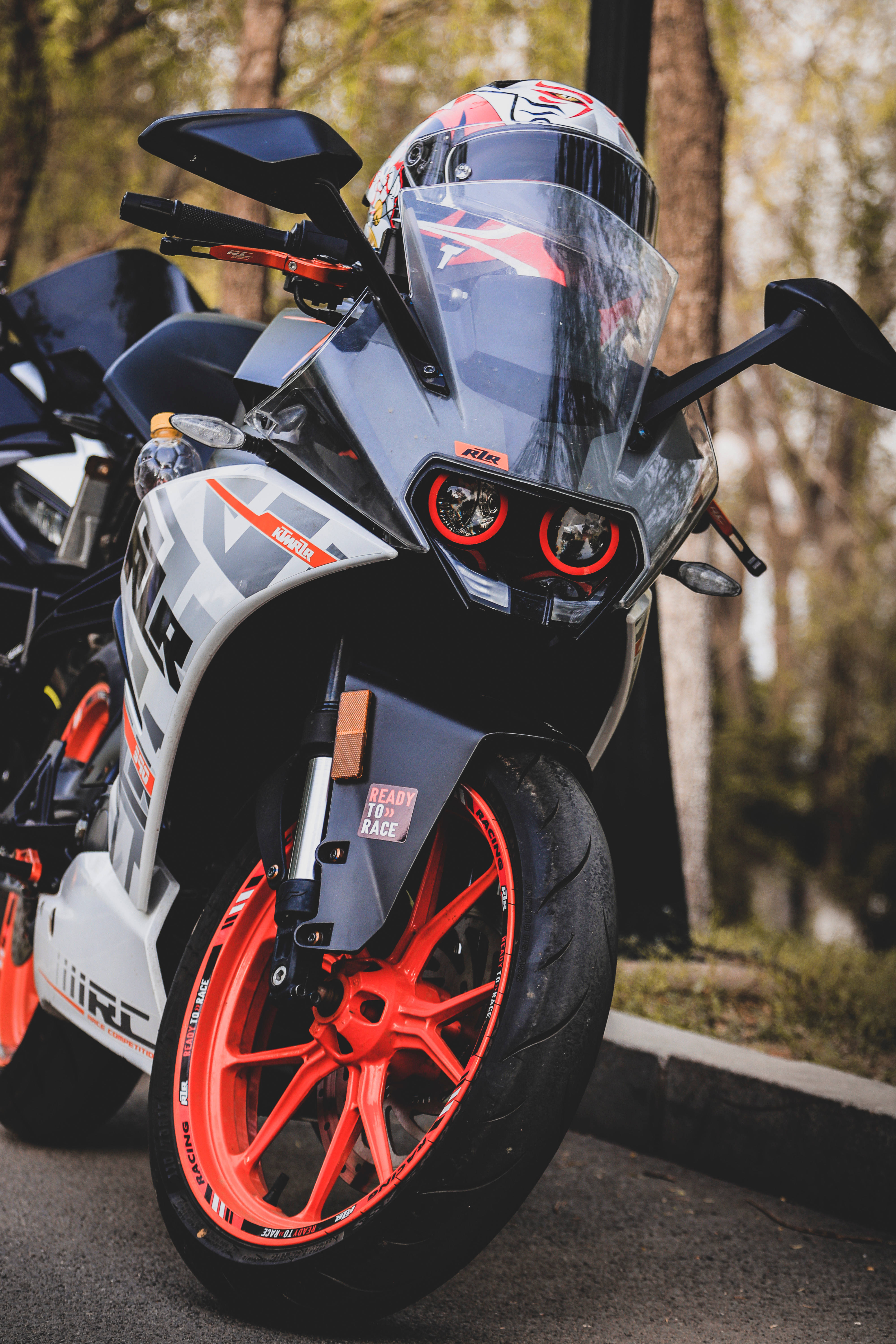 Ktm Rc 390 桌布 壁紙 — 一輛時尚的KTM運動機車，配備鮮紅色車輪和騎士安全帽，停放在綠樹成蔭的街道上。