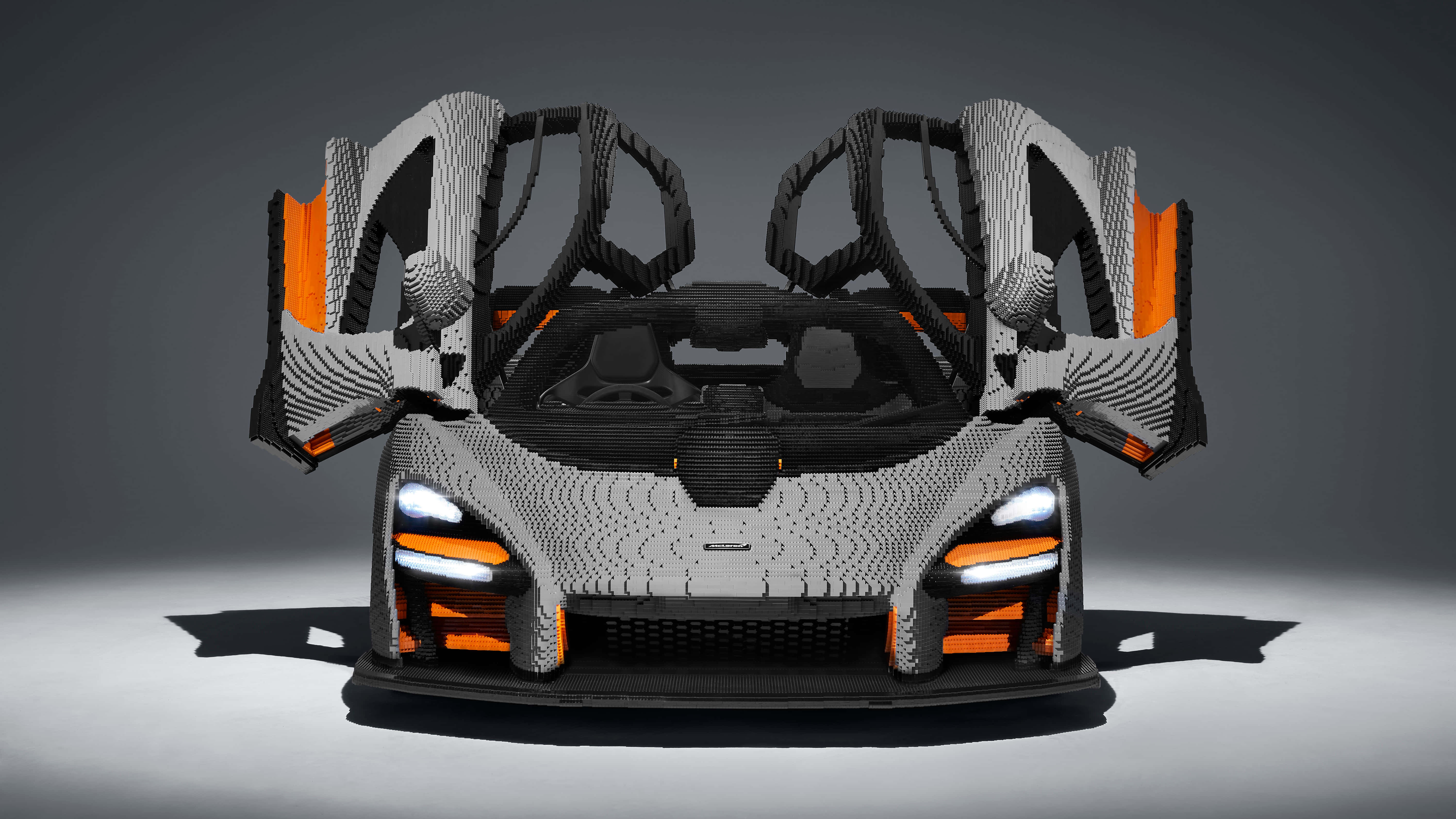 Cool Mclaren 桌布 壁紙 — 一輛時尚的McLaren跑車完全由LEGO積木建造，帶有橙色點綴和打開的蝴蝶門，背景為灰色工作室。