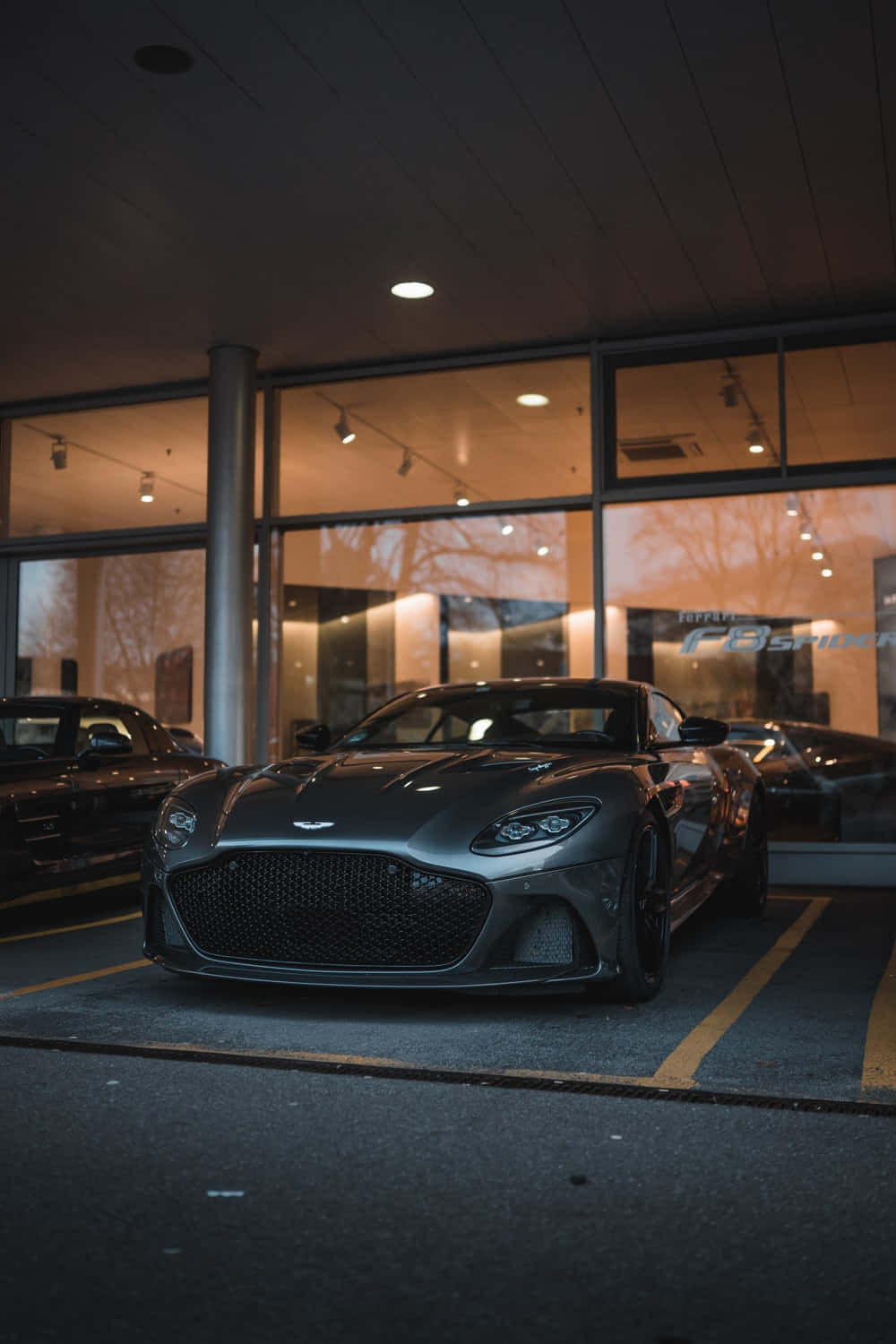 奧斯頓馬丁 桌布 壁紙 — 一輛時尚的啞光灰色 Aston Martin Vantage 在夜間停放在現代玻璃天篷下，溫暖的車內燈光反射在其拋光表面上。