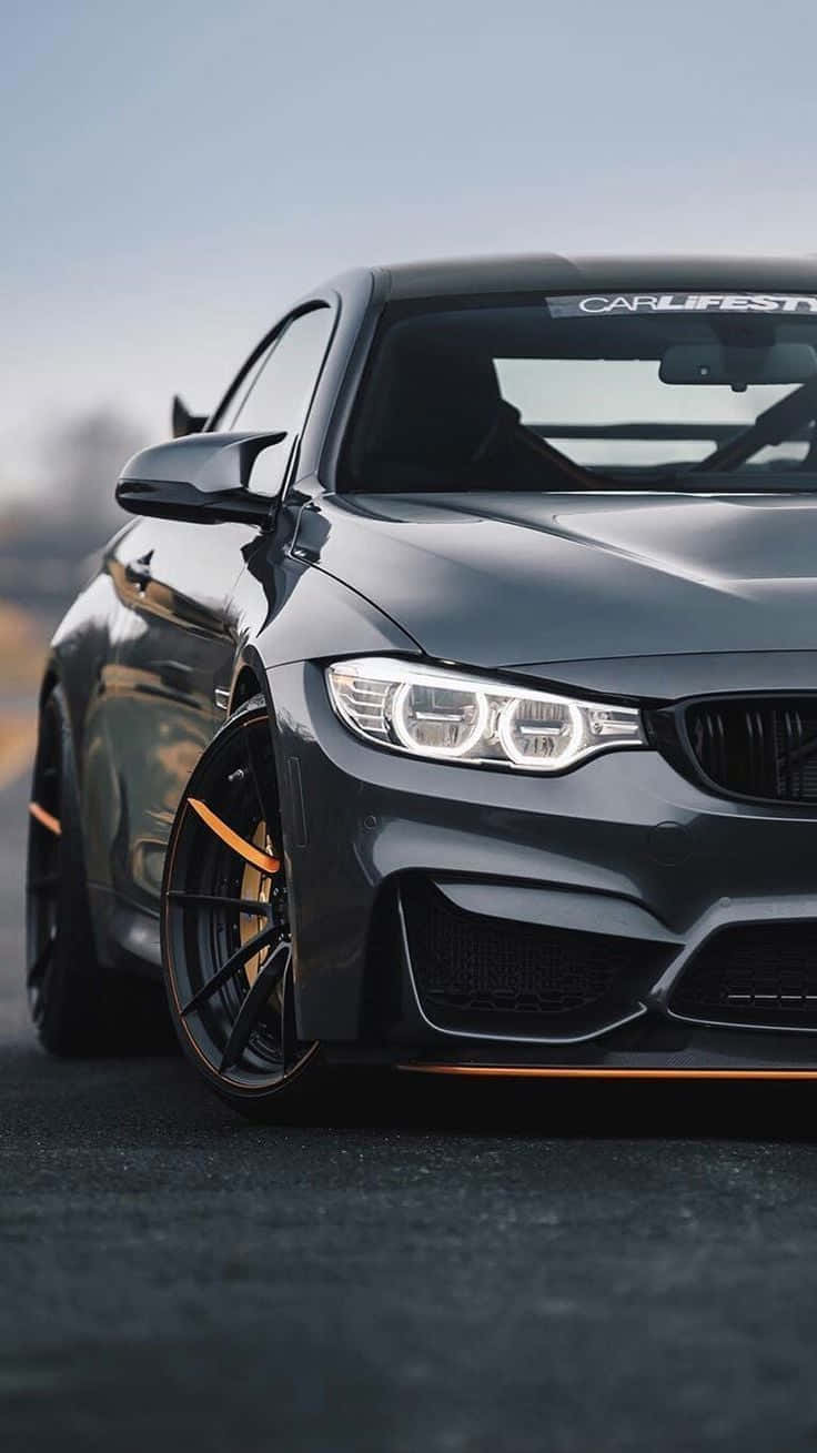 BMW 動圖 桌布 壁紙 — 一輛時尚的啞光灰色BMW M4，帶有橙色點綴和發光的車頭燈，停在瀝青路面上，處於柔和日光下。