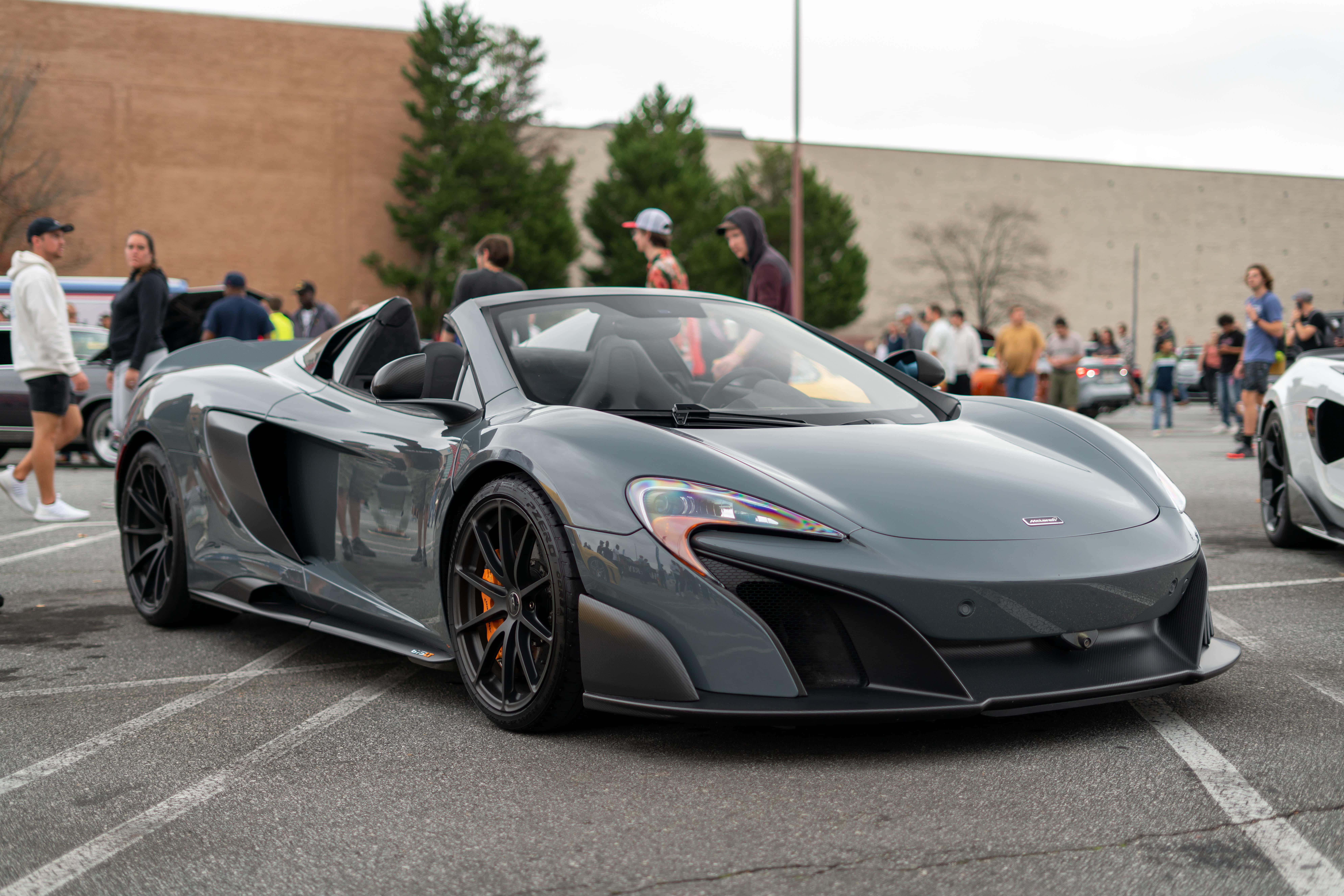 Mclaren 675lt Spider 桌布 壁紙 — 一輛時尚的啞光灰色McLaren跑車停放在停車場，人們在陰天欣賞它。