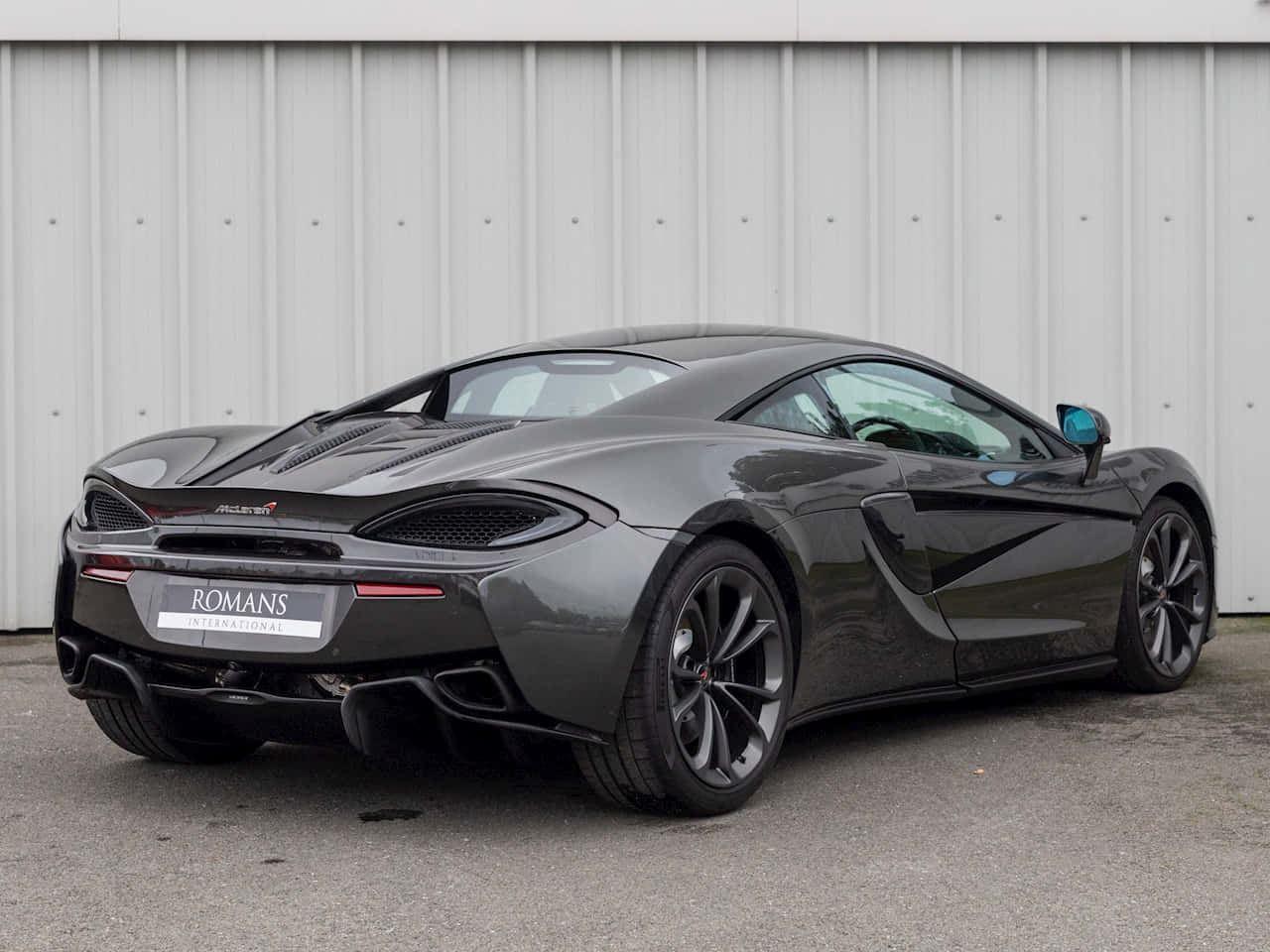 Mclaren 540c 桌布 壁紙 — 一輛時尚的啞光灰色McLaren 600LT跑車停靠在波紋金屬牆旁，車牌上寫著ROMANS INTERNATIONAL。
