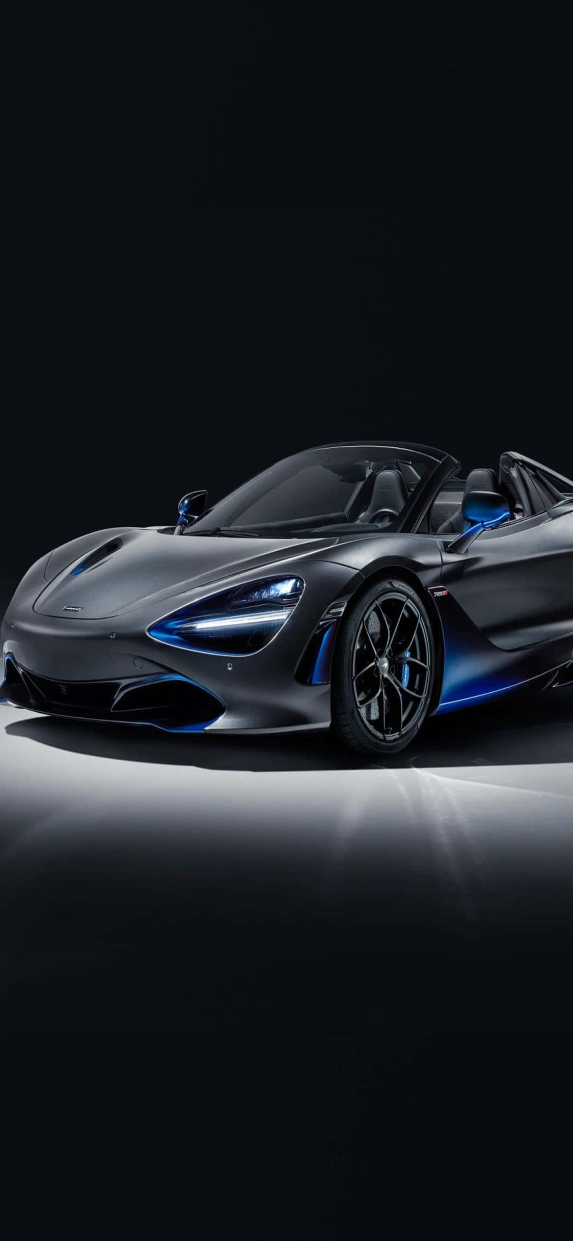 iPhone Xs Mclaren 720s 桌布 壁紙 — 一輛時尚的啞光灰色McLaren 720S Spider，帶有發光的藍色飾邊和車頭燈，在黑色工作室背景下亮起。