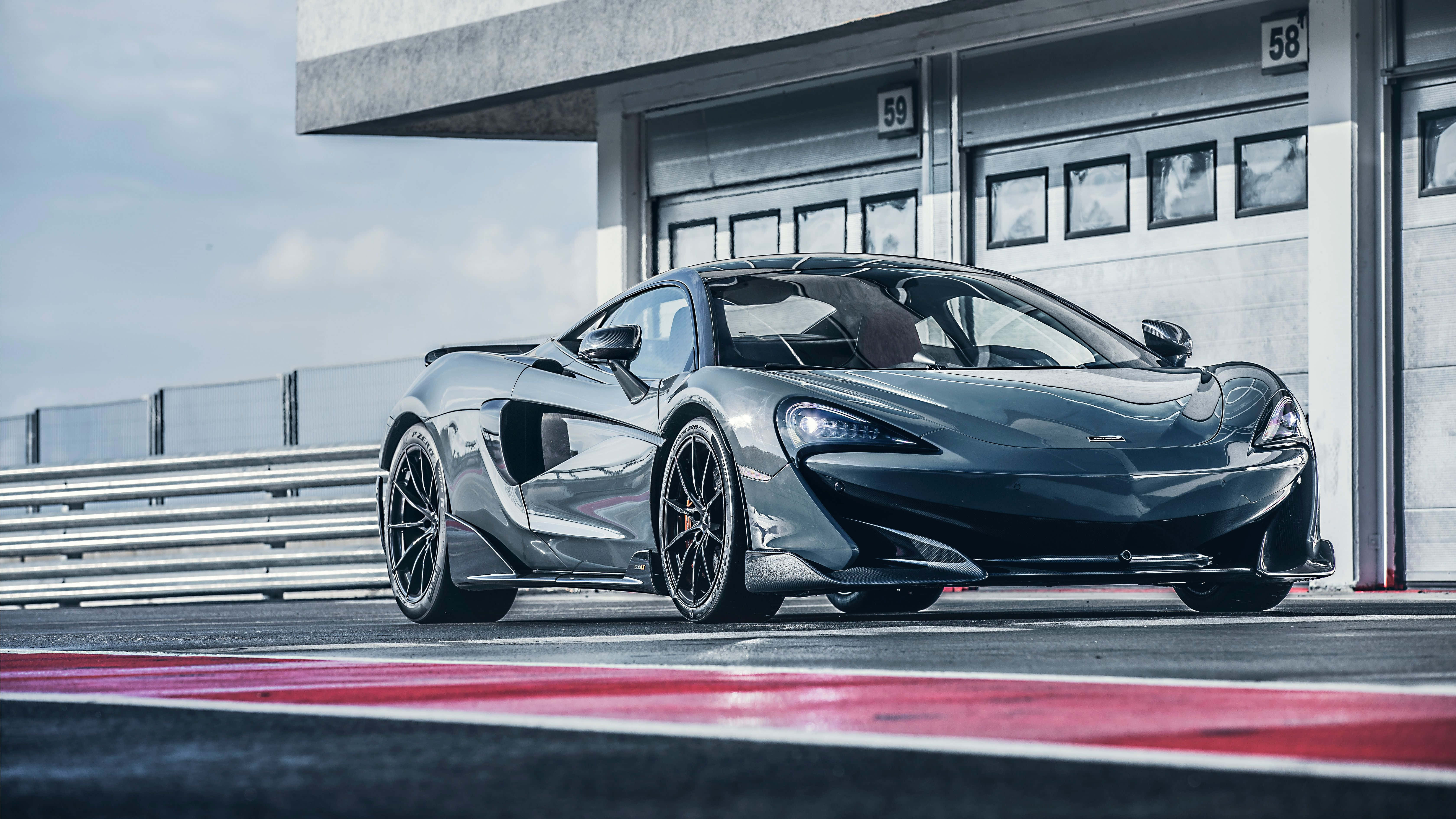 Mclaren 600lt 桌布 壁紙 — 一輛時尚的啞光灰色McLaren跑車停泊在賽道上，旁邊是帶有紅色路緣石的混凝土車庫，天空晴朗。