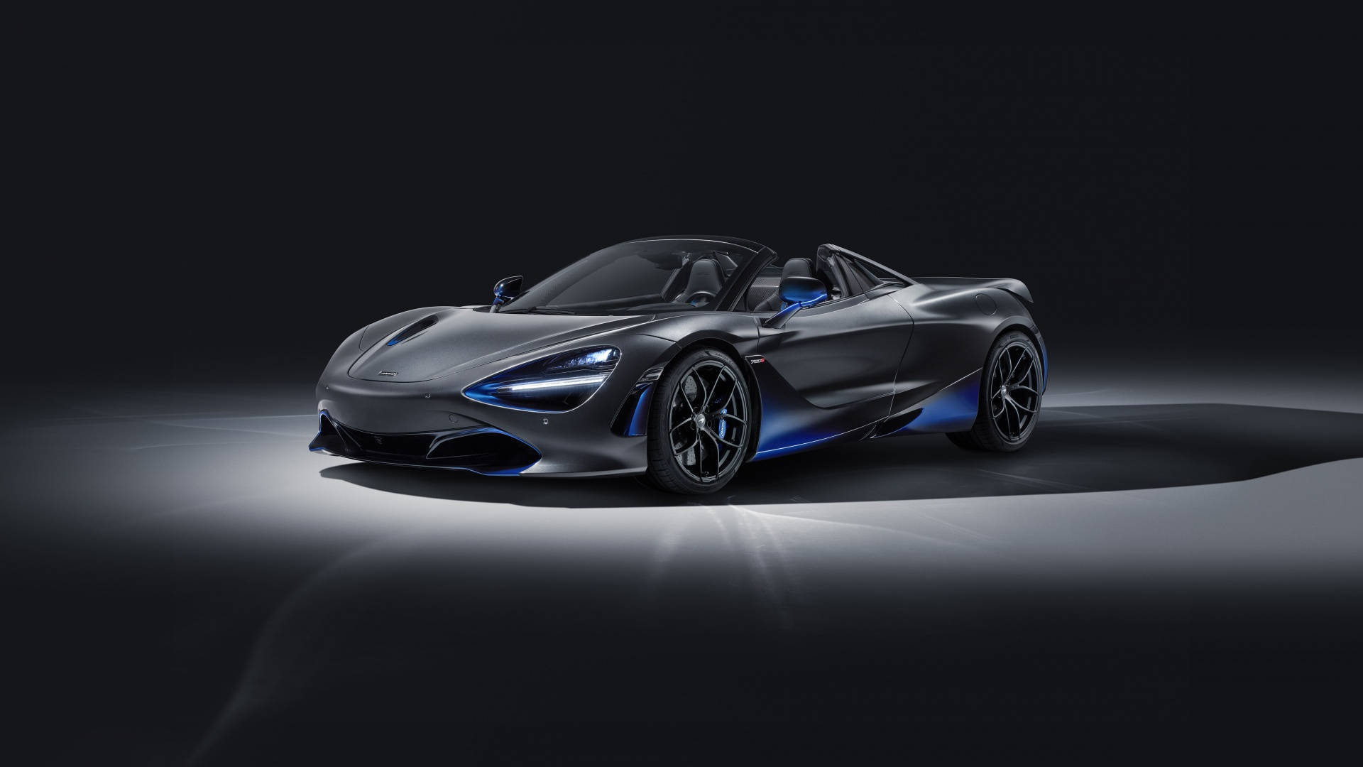 Mojave 桌布 壁紙 — 一輛時尚的啞光灰色McLaren 720S Spider，帶有發光的藍色點綴，在黑暗的攝影棚背景下戲劇性地照亮。